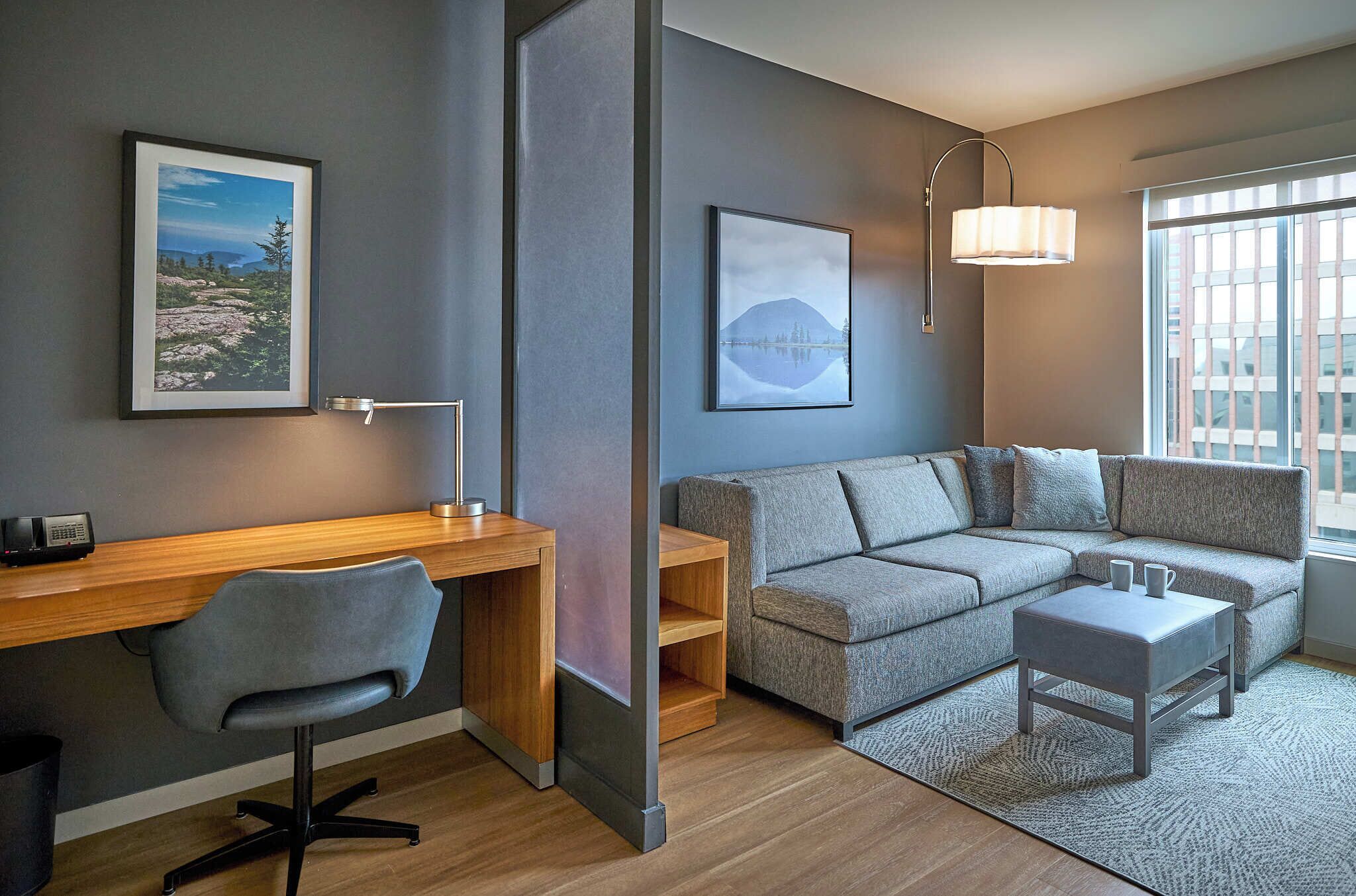 A modern room with a desk, chair, sofa, and ottoman. There are two framed pictures on the wall, a lamp, and a window with blinds.