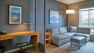 A modern room with a desk, chair, sofa, and ottoman. There are two framed pictures on the wall, a lamp, and a window with blinds.