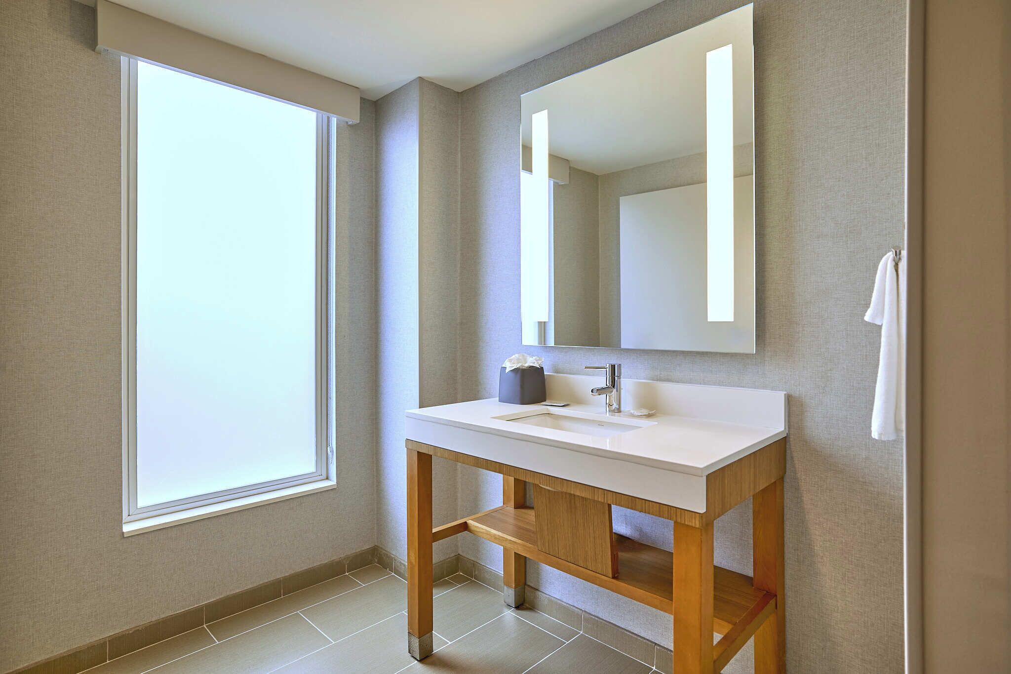 A bathroom with a large window, a white sink, a mirror, and a towel hanging on the wall.