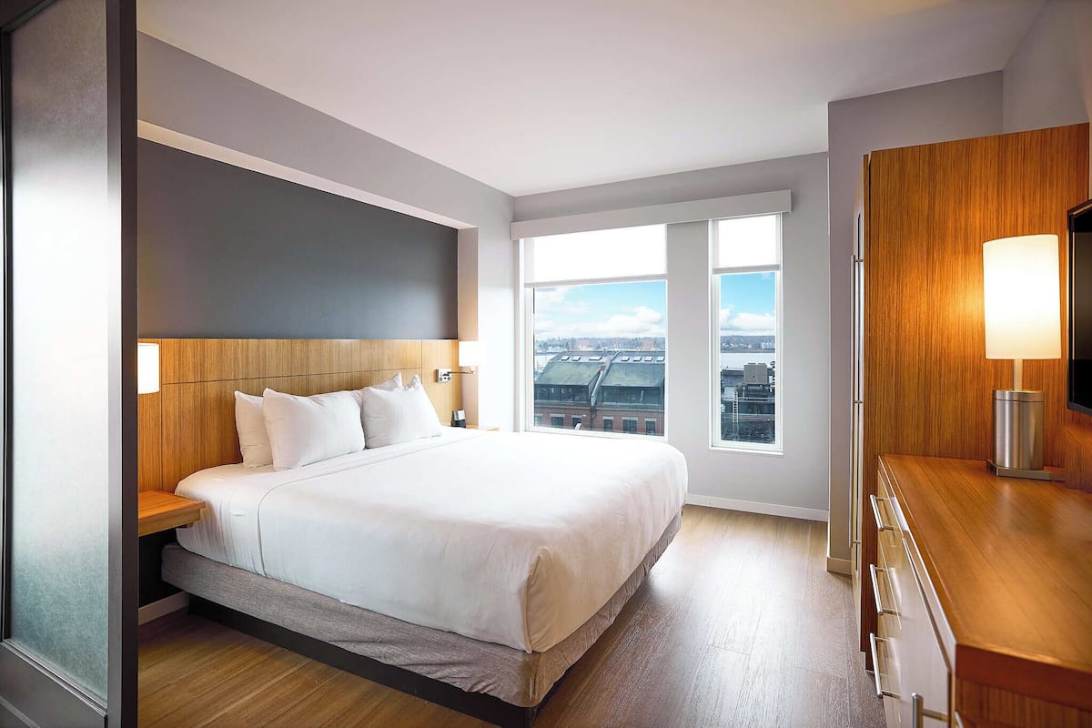 A modern hotel room with a large bed, a wooden headboard, a wooden desk, a nightstand, a TV, and a window with a city view.
