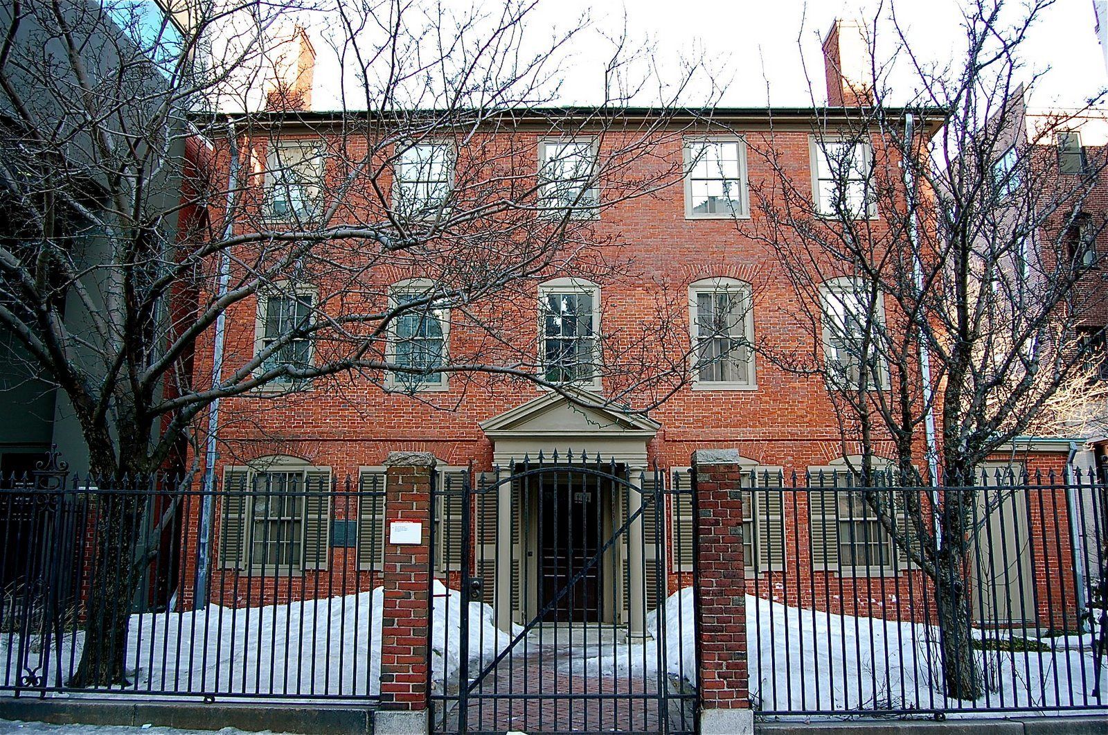 A red brick building with a symmetrical facade, a central entrance with a pediment, and a black metal fence surrounding the property.