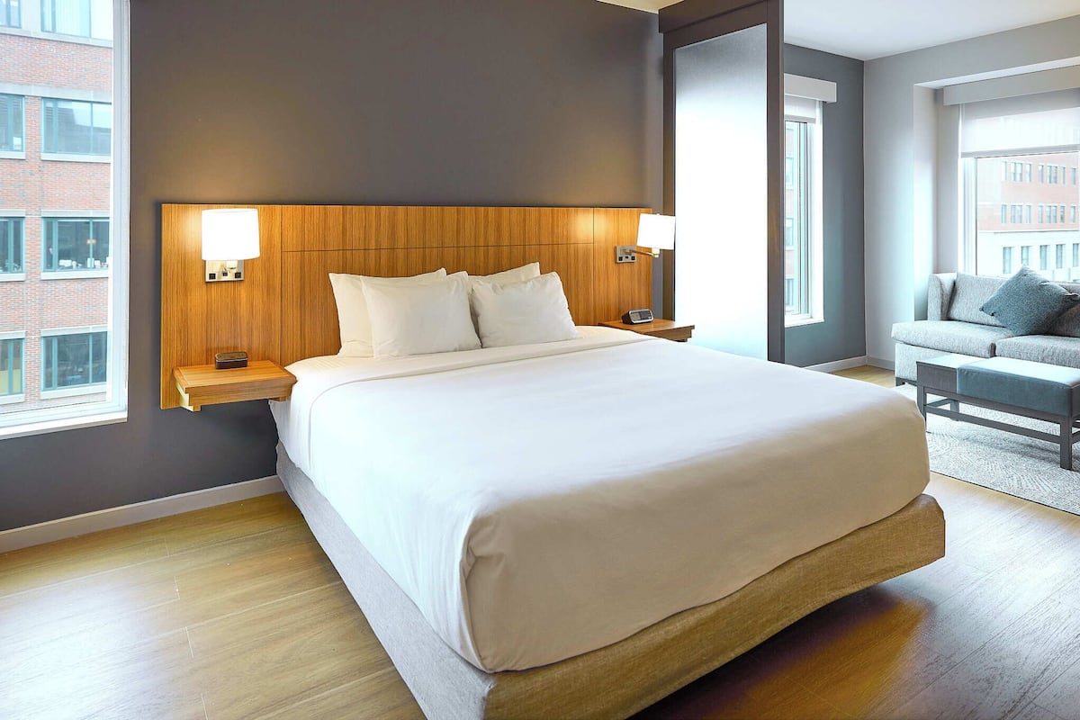 A hotel room with a large bed, two bedside tables with lamps, a grey sofa, and a view of a brick building.