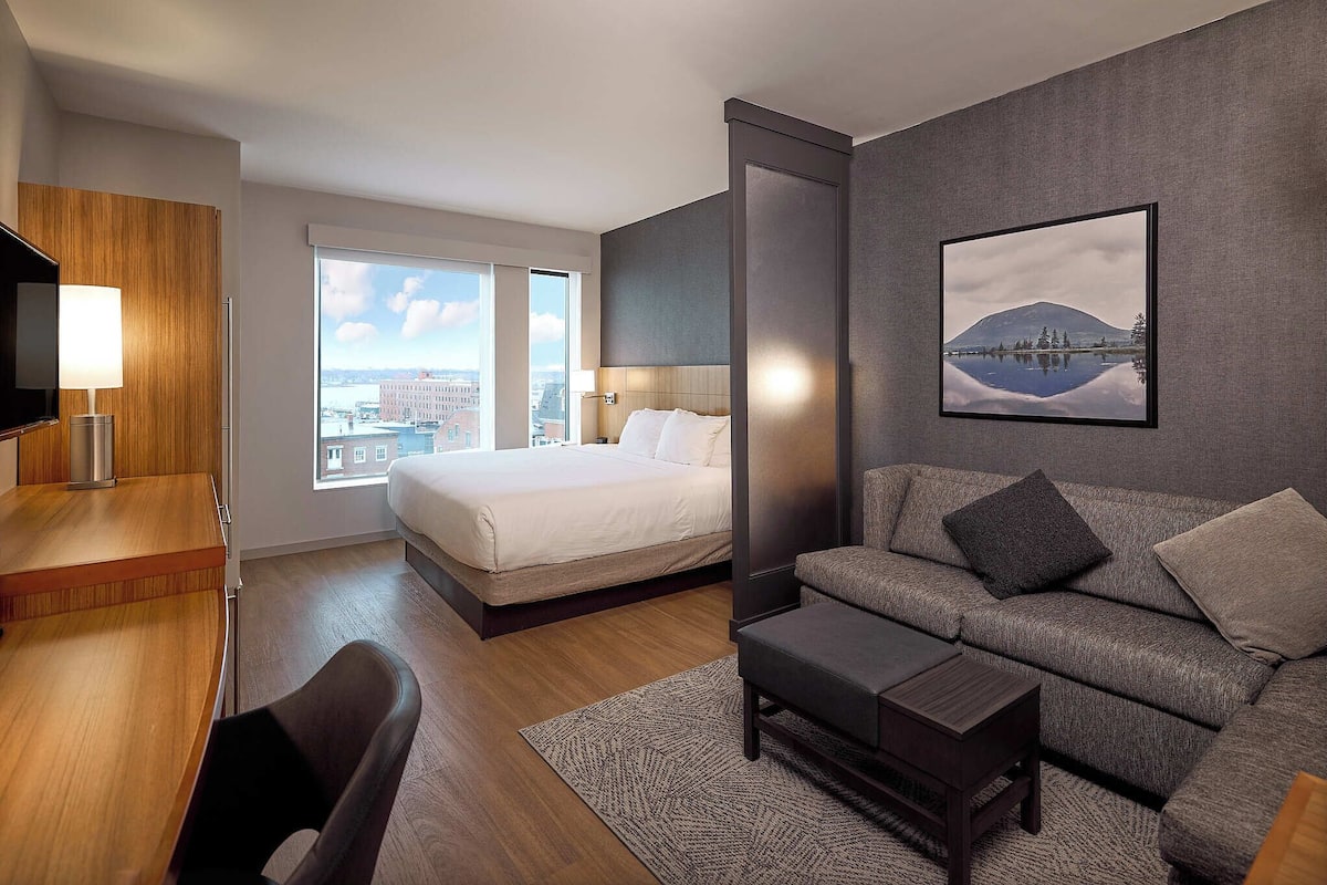 A modern hotel room with a bed, sofa, desk, and a large window offering a city view.