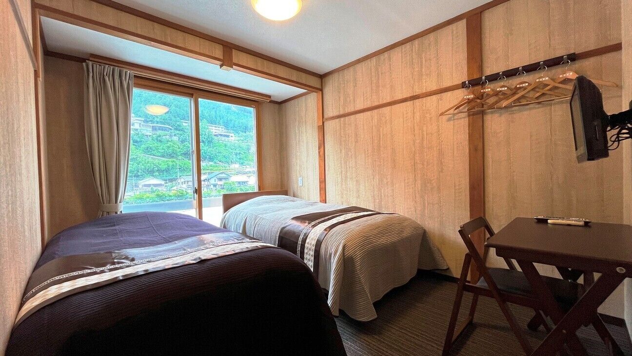 Et hotelværelse med to senge, et bord og en stol. Et vindue med gardiner og udsigt til grøn bevoksning udenfor.