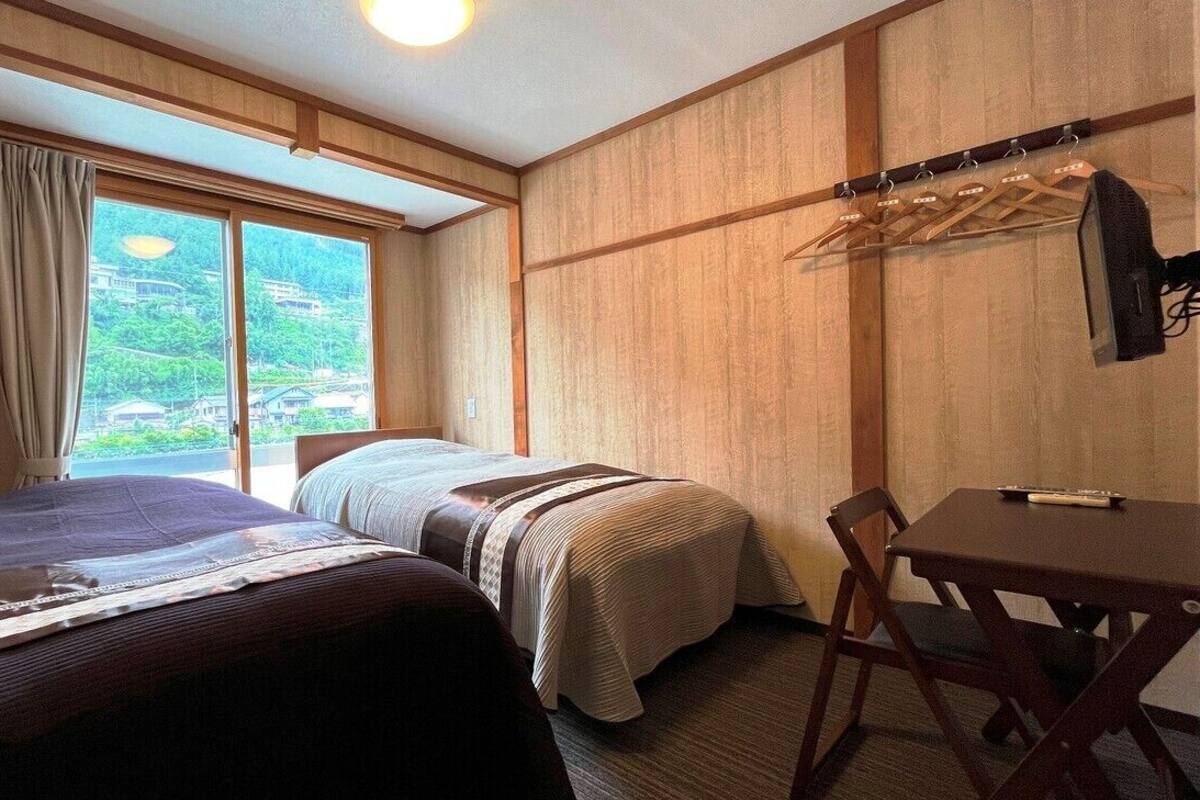 A hotel room with two beds, a table, and a chair. There is a window with curtains and a view of greenery outside.