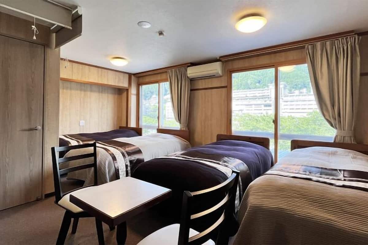A room with two beds, a table, and a chair. There are windows with curtains, a ceiling light, and an air conditioning unit.
