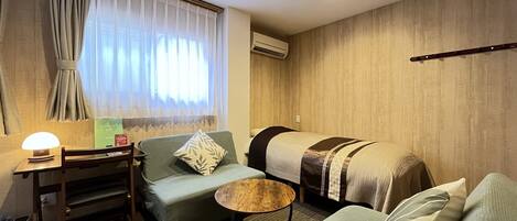 A hotel room with a green sofa, a wooden table with a lamp, a small round table, and a bed with a striped blanket.