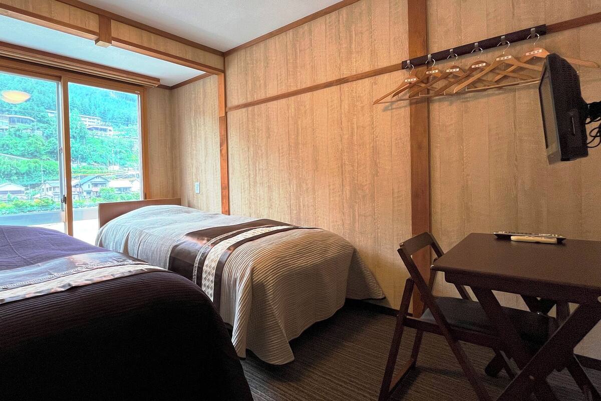 A hotel room with two beds, a table, and a chair. There is a window with curtains, a coat rack, and a television.