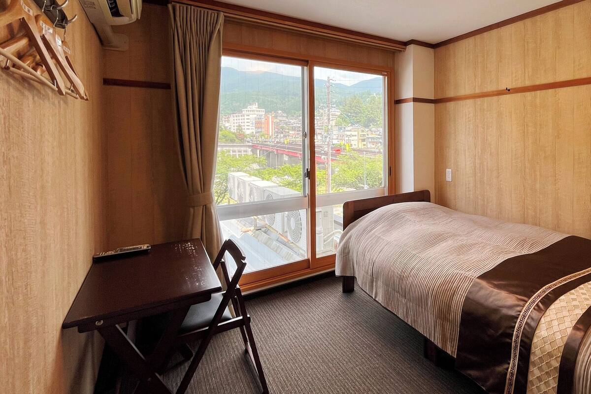 A hotel room with a bed, a desk, and a chair. There is a window with a view of buildings and a bridge.