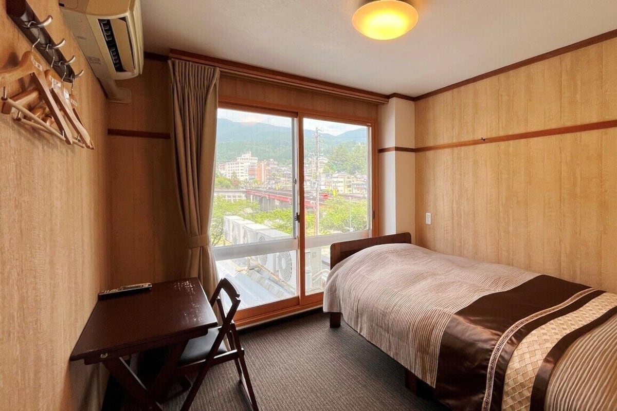 A single bed with a striped bedspread, a desk with a chair, and a large window with a view of buildings and mountains.