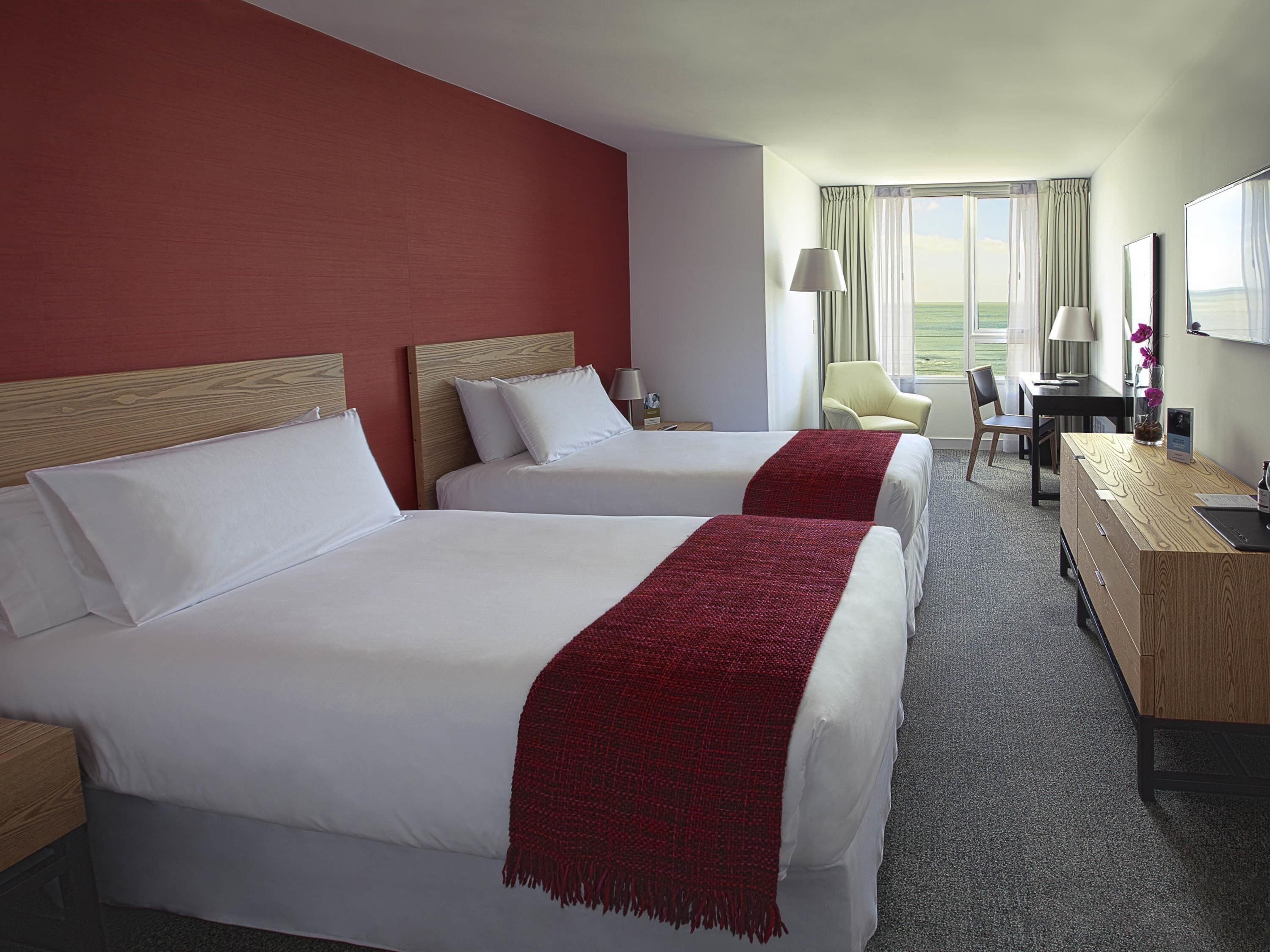A hotel room with two beds, a desk, a chair, and a view of the outdoors.