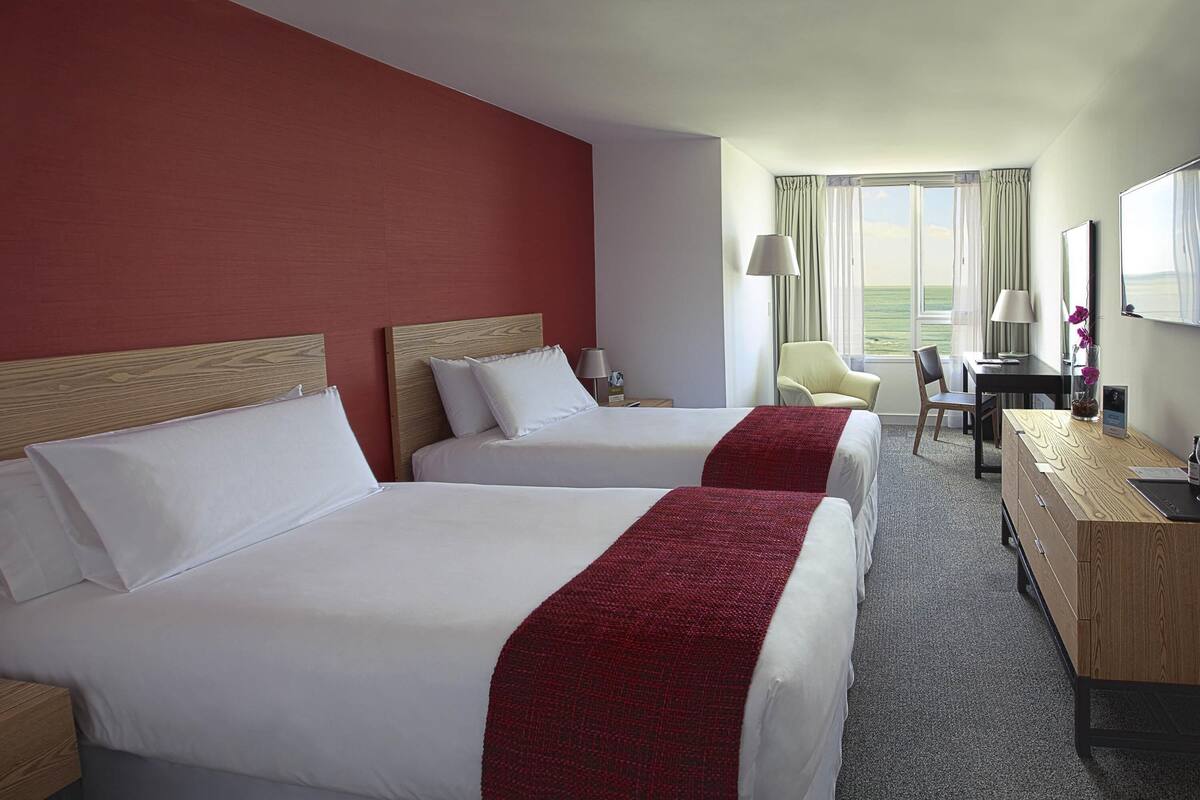 A hotel room with two beds, a desk, a chair, and a view of the outdoors.