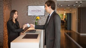 A reception desk with a monitor displaying "Mercure Hotels" and a man handing a card to a woman. - Mercure Montevideo Punta Carretas (Montevideo)