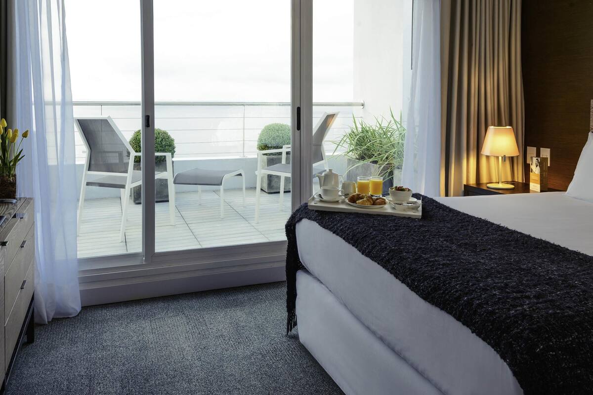 A hotel room with a bed, a tray with breakfast items, a nightstand with a lamp, and a view of the balcony with outdoor furniture.