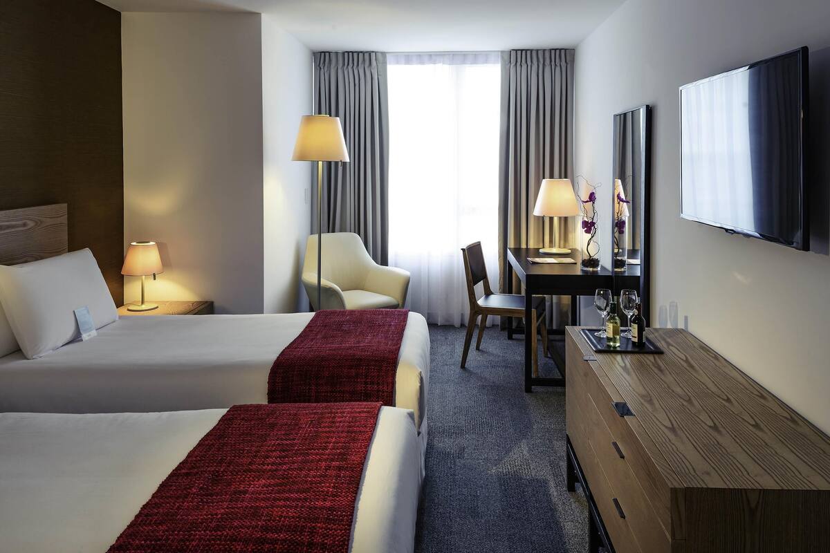 A hotel room with two beds, a desk, a chair, a TV, and a window with curtains.