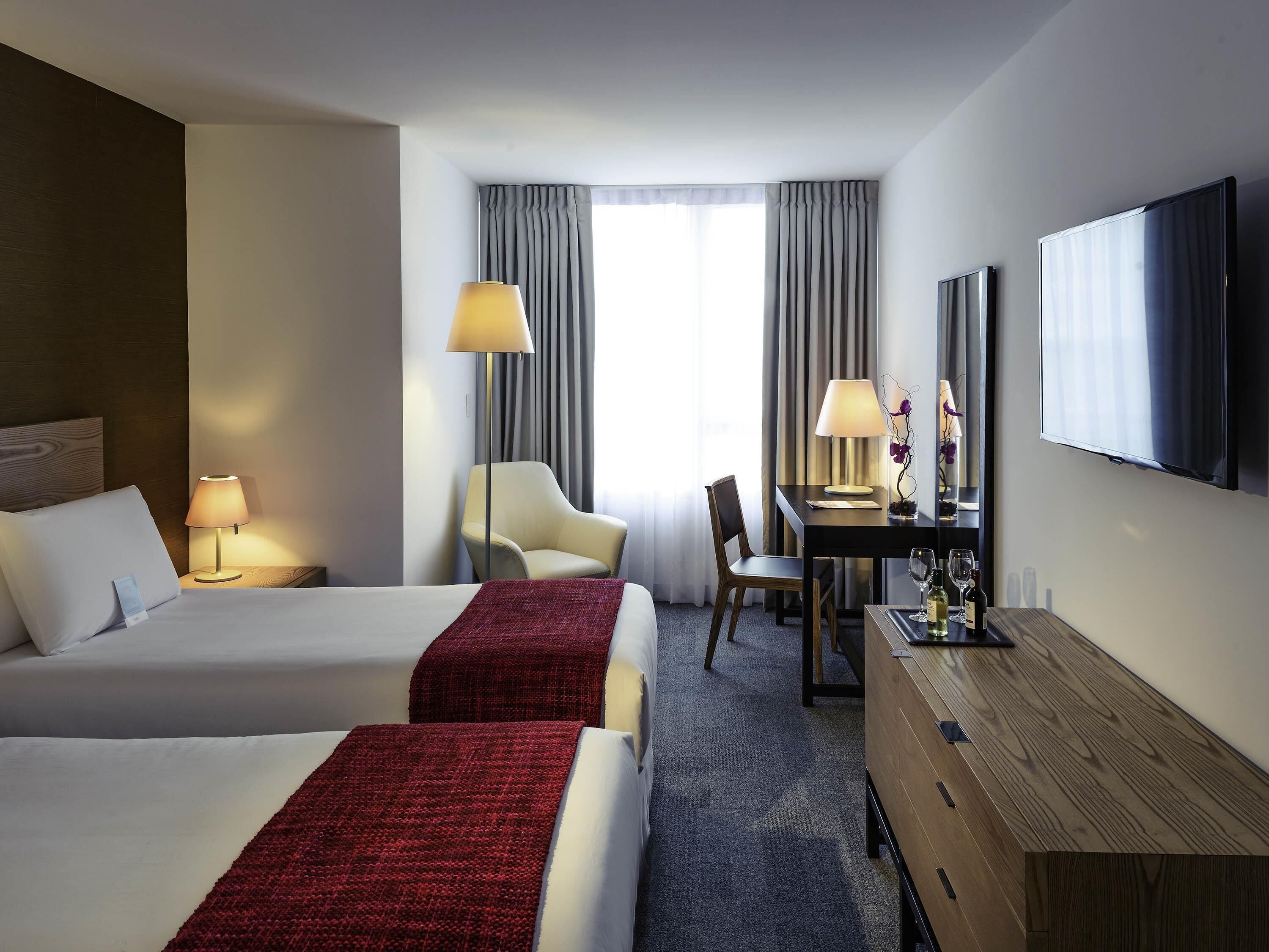 Habitación de hotel con dos camas, un escritorio con lámpara, una silla, un televisor y un ventanal con cortinas.