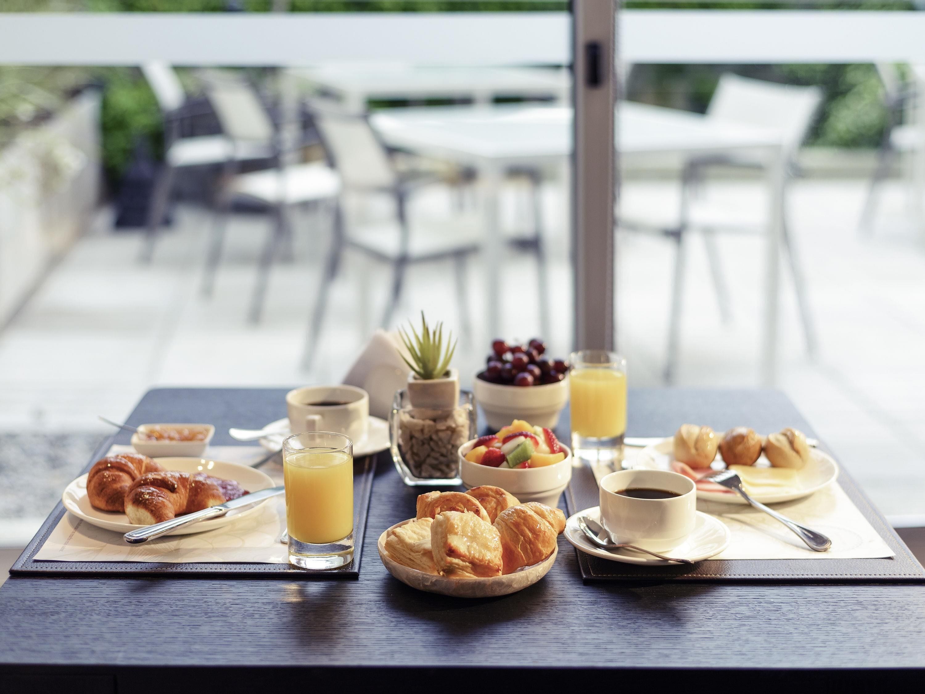 A breakfast setting with croissants, fruit, juice, and coffee on a table with a view of outdoor seating.