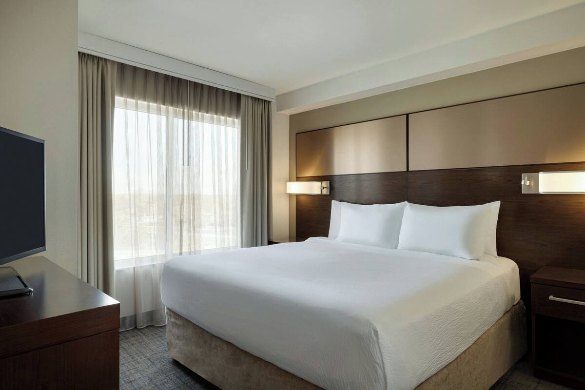 A hotel room with a large bed, a desk with a computer, a TV, and a window with curtains.