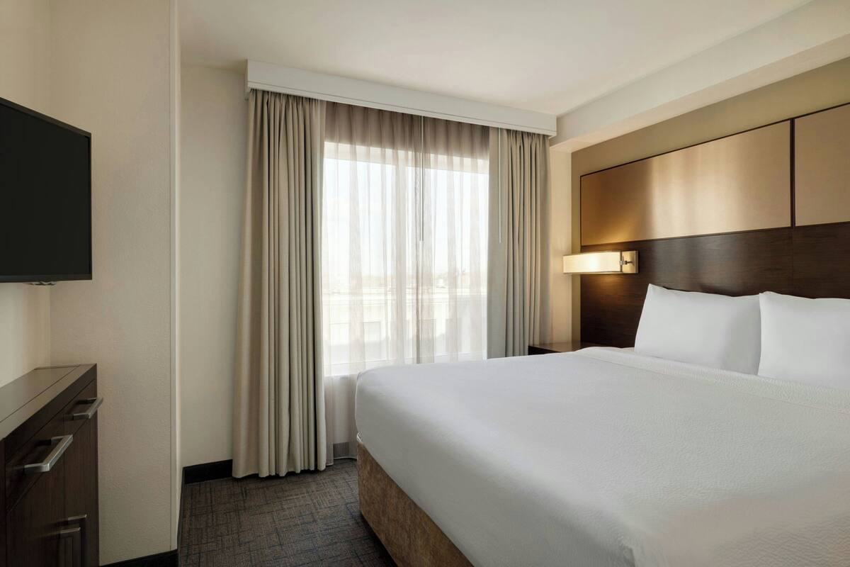 A hotel room with a large bed, a flat-screen TV, a desk with drawers, and a window with curtains.