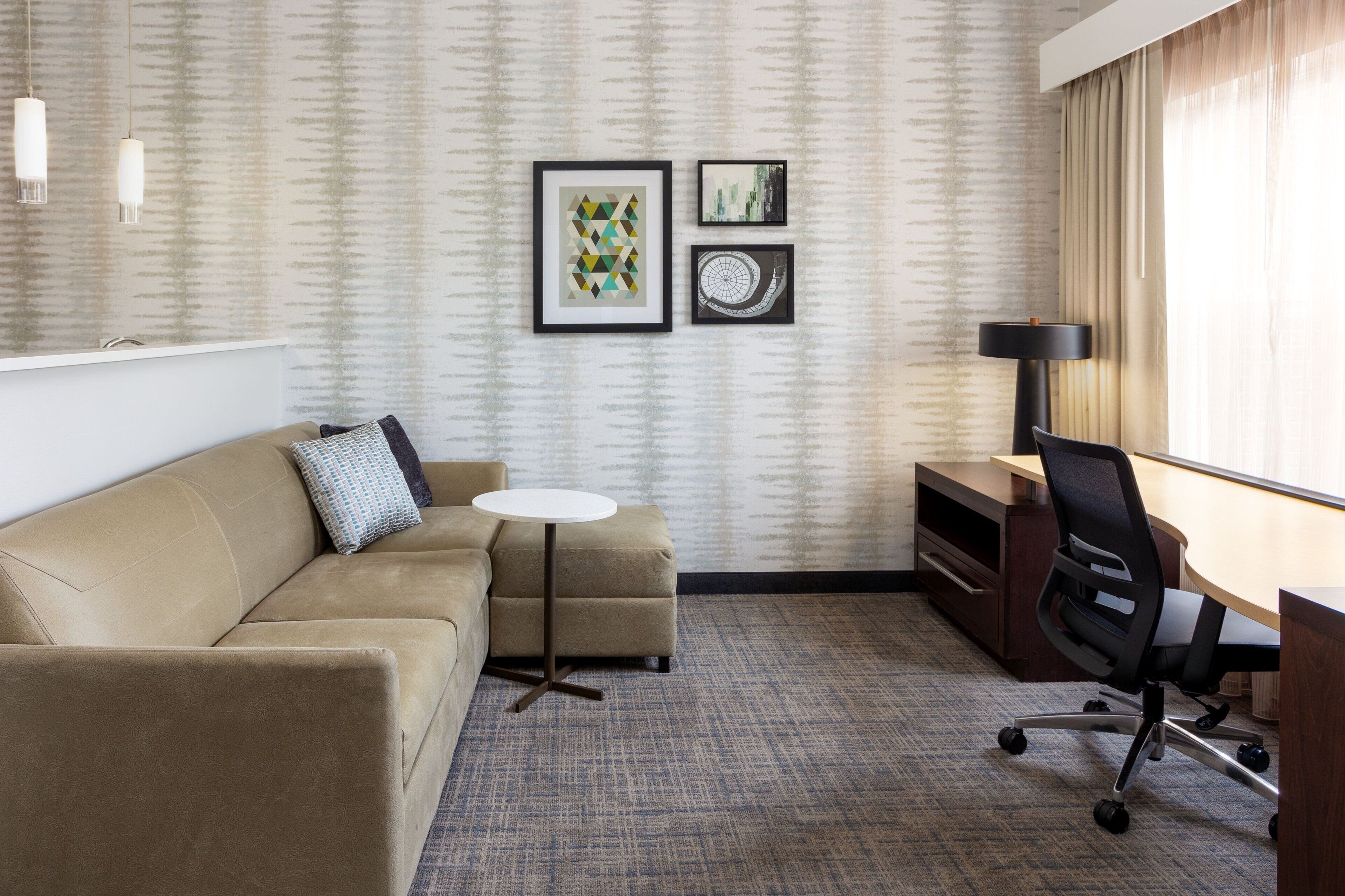 Une chambre d’hôtel avec un canapé beige, une petite table ronde, un bureau avec une chaise et deux œuvres d’art encadrées accrochées au mur.