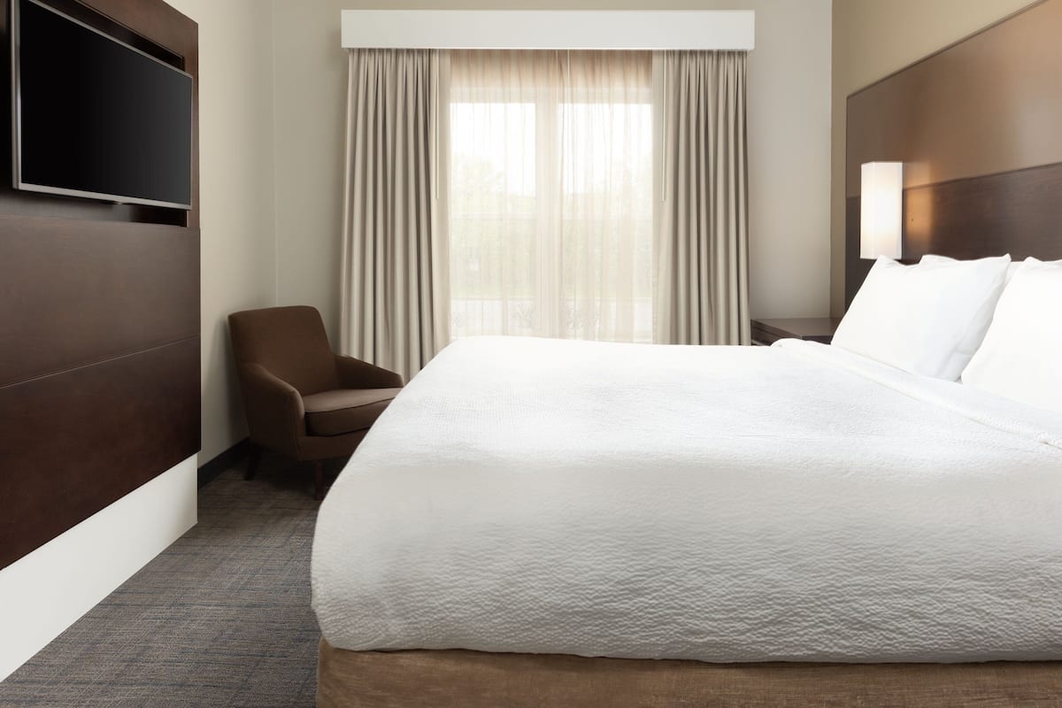 A hotel room with a large bed, a flat-screen TV mounted on the wall, a chair, a window with curtains, and a nightstand with a lamp.