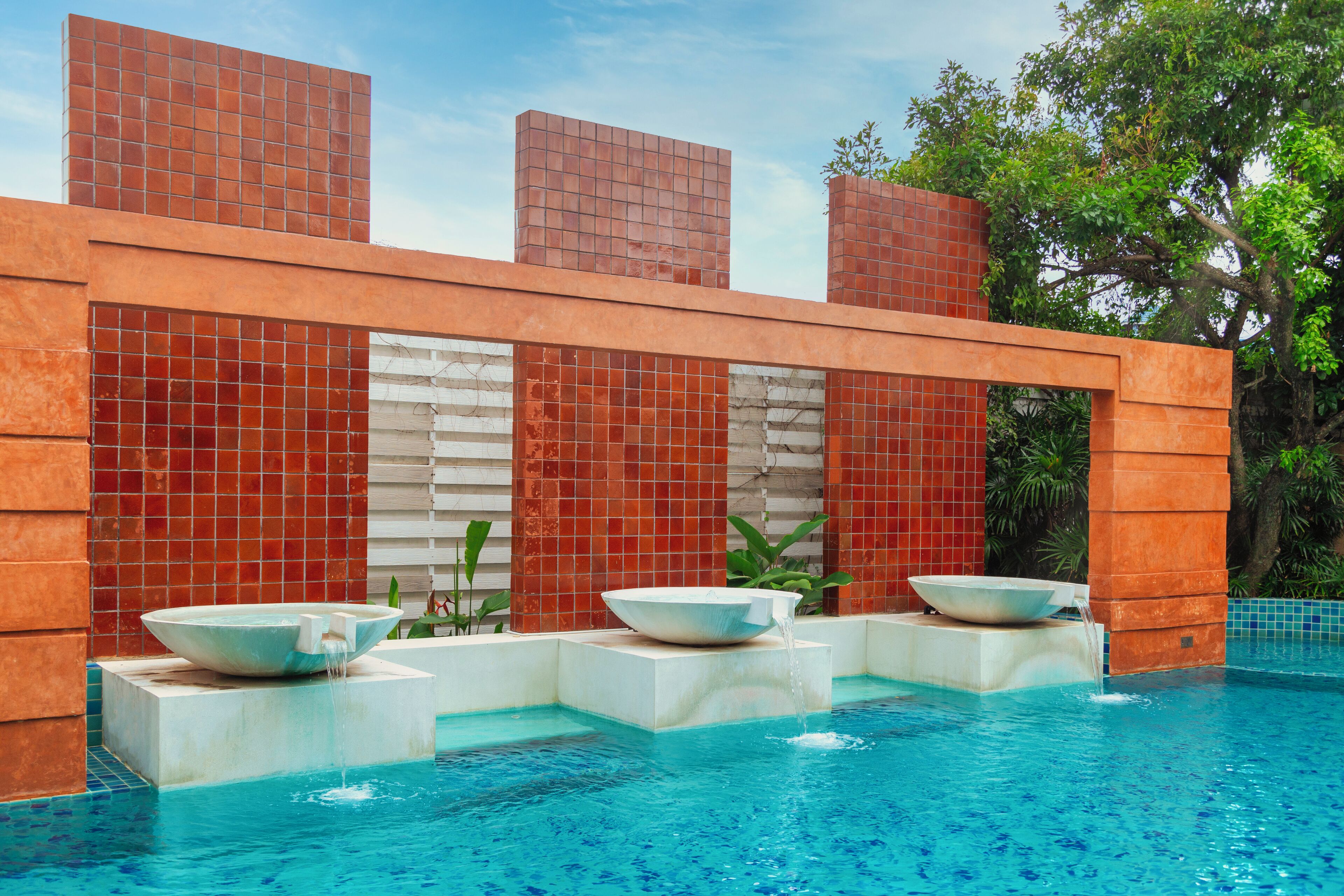 Un área de piscina moderna con diseño geométrico, que presenta una fuente central y una pared de azulejos.