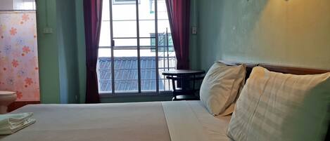 A hotel room with a bed, a table, a lamp, a view of the outside, and a toilet.