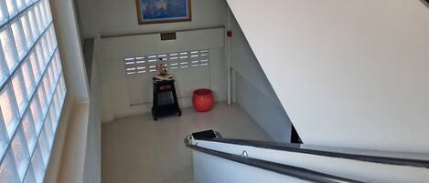 Uma escadaria com listras azuis leva a um patamar. Há uma moldura com um quadro na parede acima do patamar, uma pequena mesa preta com um vaso vermelho e uma placa sobre a mesa.