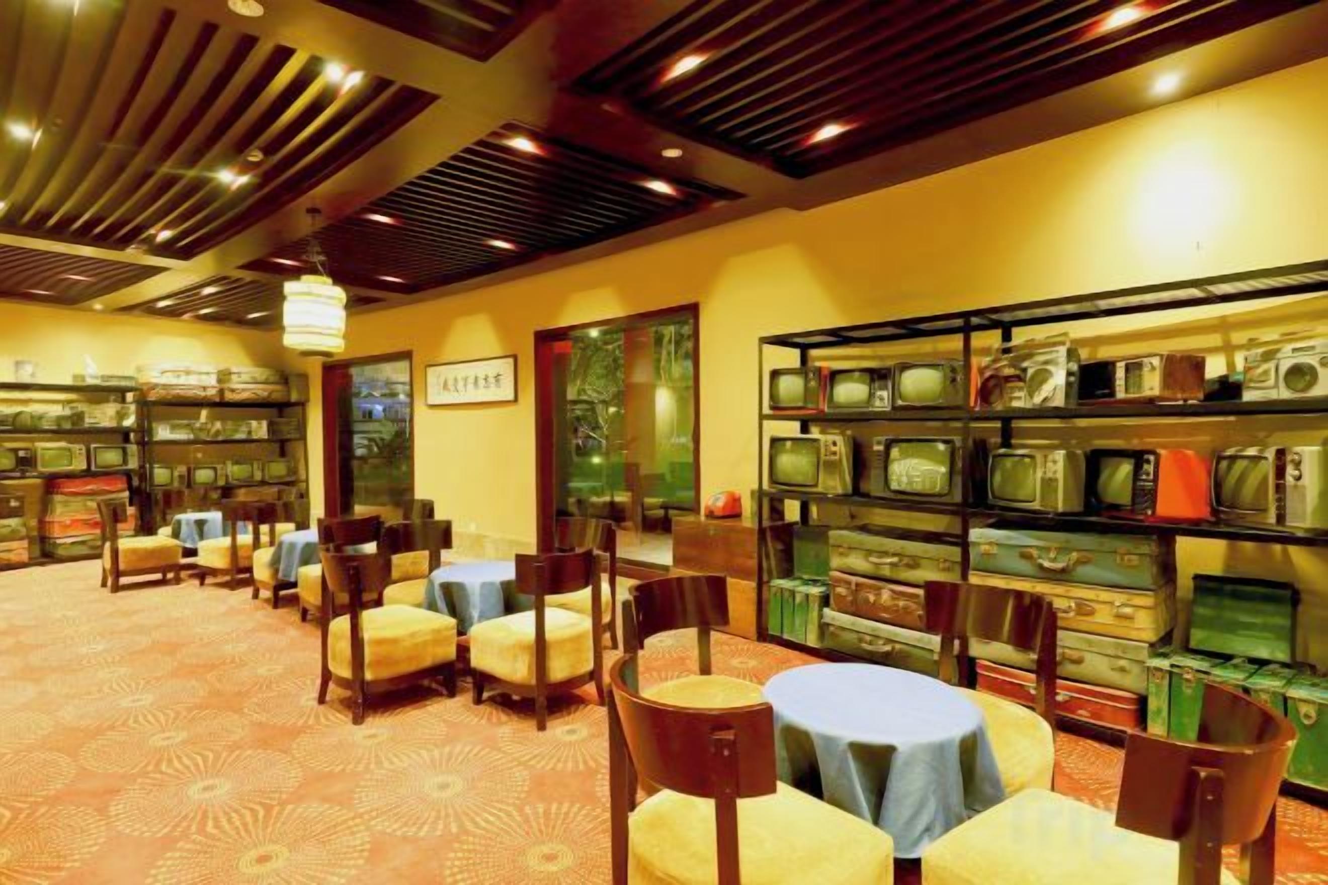 A room with multiple tables and chairs, a bookshelf with various items, and a patterned carpet.