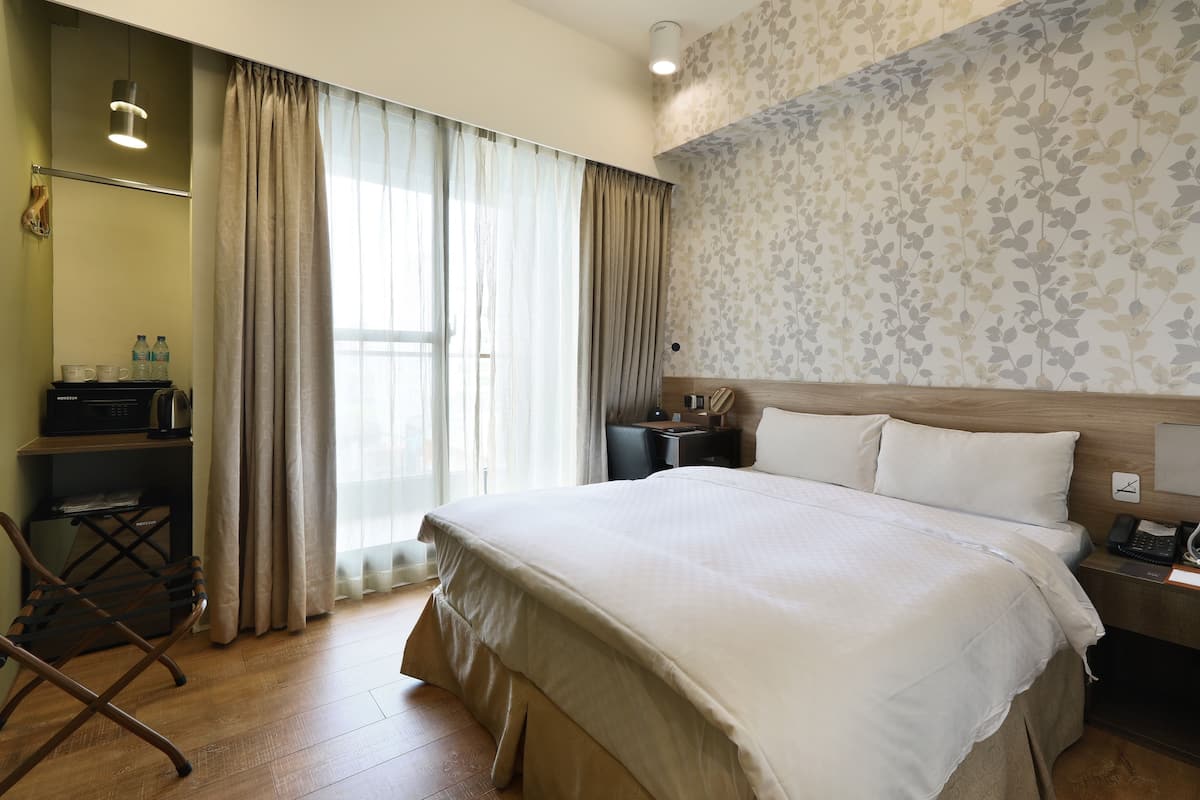 A hotel room with a large bed, a desk with a lamp, a chair, and a wardrobe.