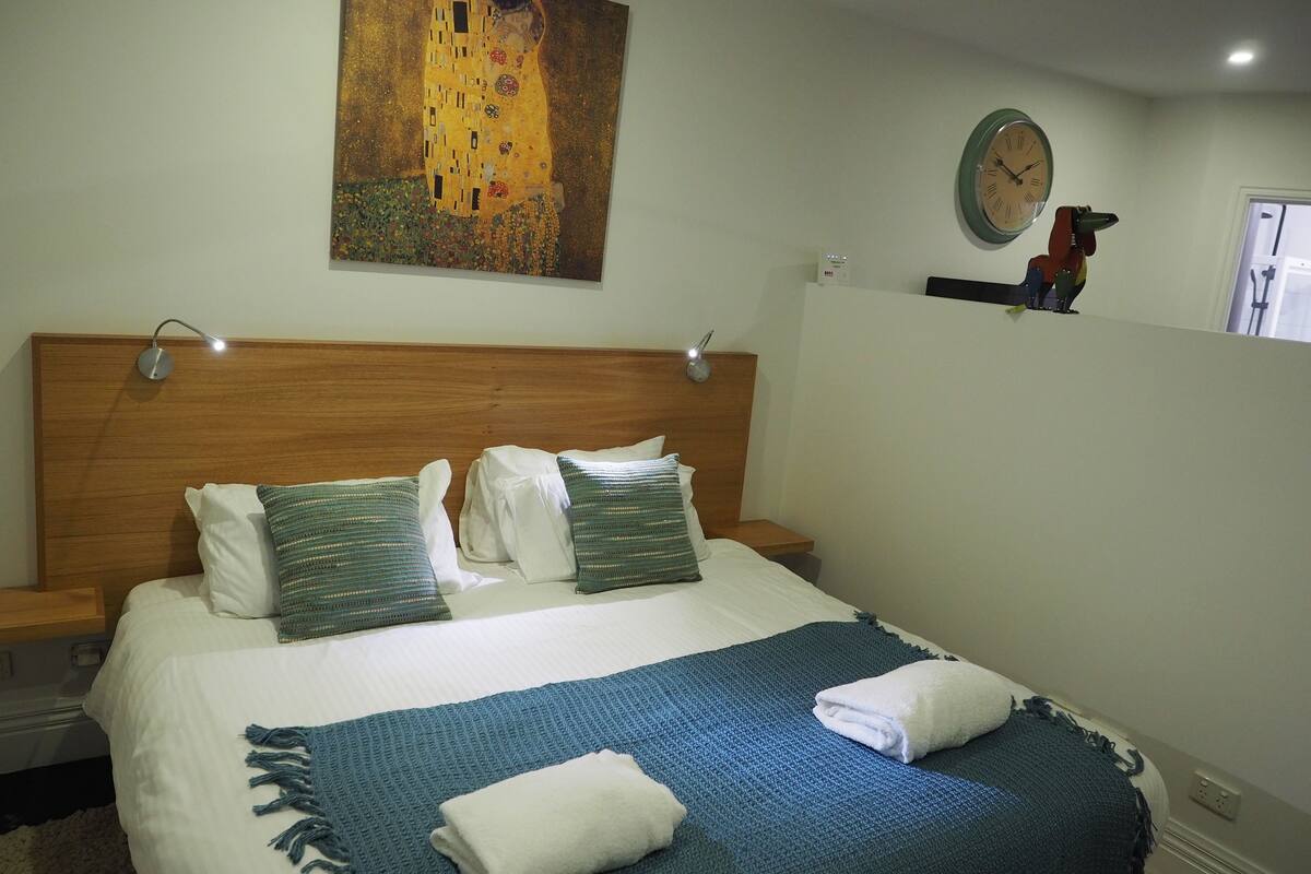 Una cama bien hecha con una manta azul, almohadas blancas y dos toallas dobladas. Un cuadro de una pareja está en la pared sobre la cama.