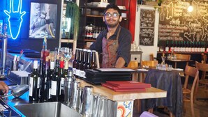Zona de bar con un bartender, botellas de vino, vasos y un menú en una pizarra.