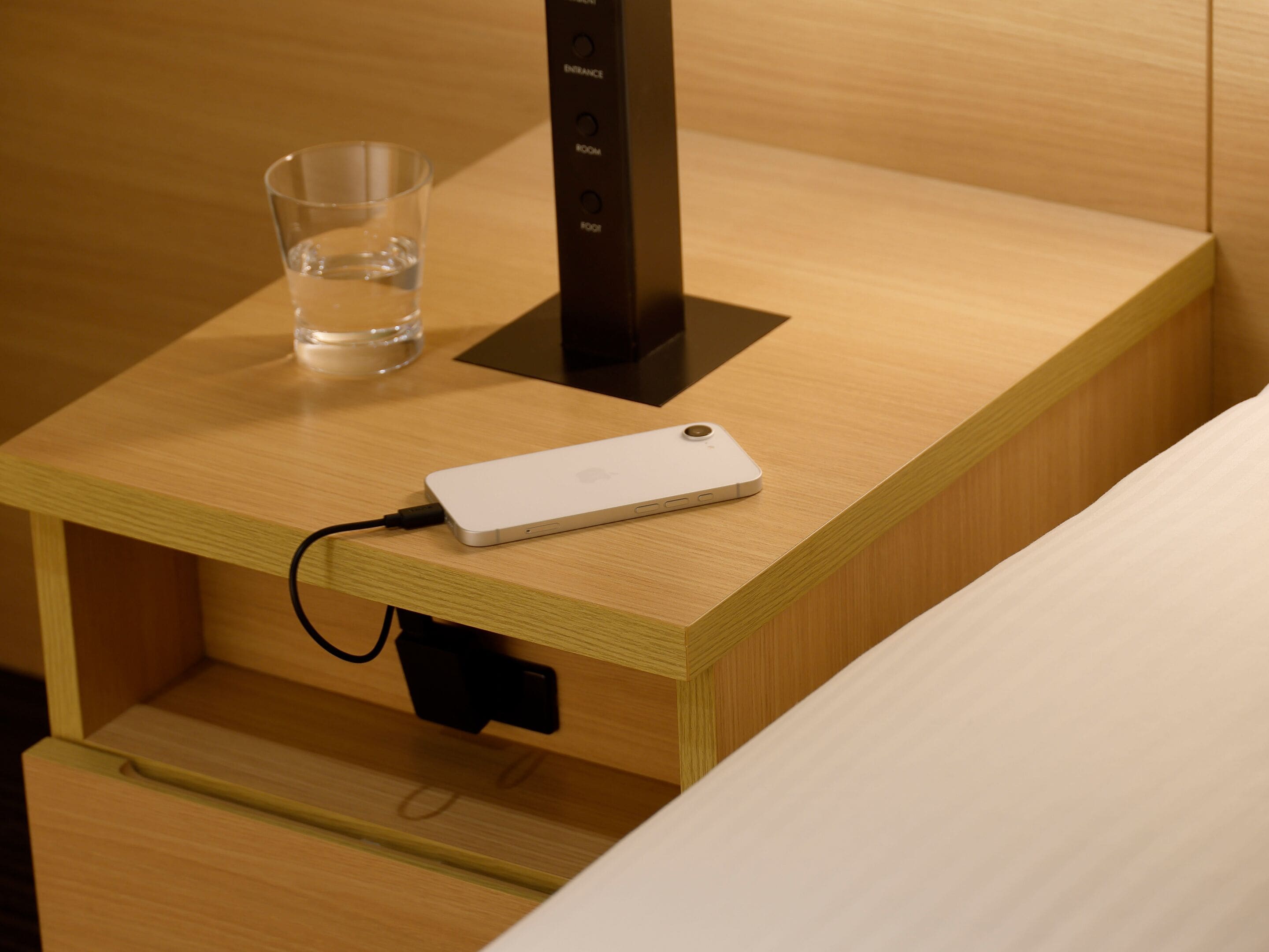 A wooden bedside table with a white electronic device and a glass of water.