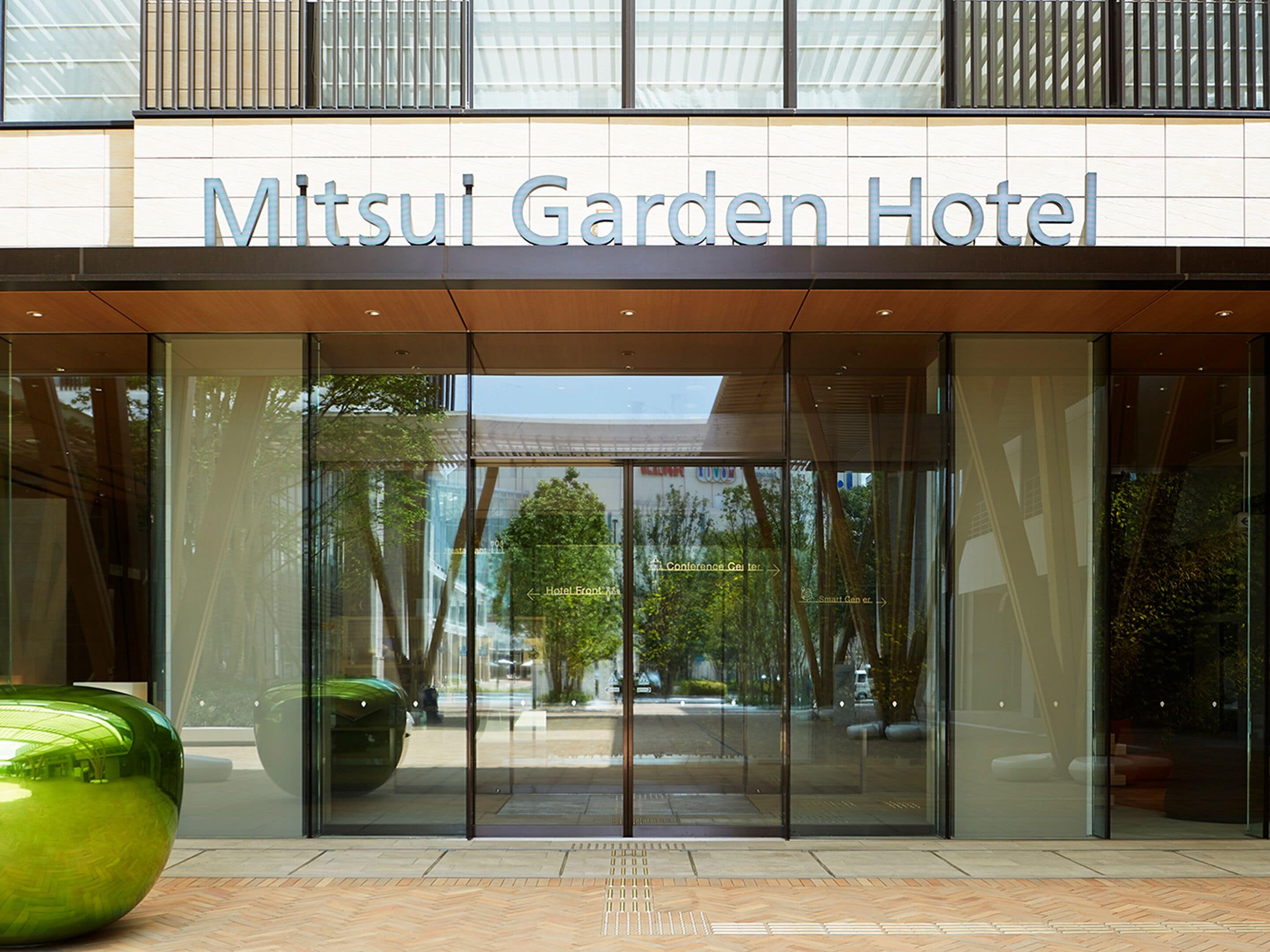 La entrada del Mitsui Garden Hotel, con fachada de vidrio transparente, un gran macetero verde y señalización direccional hacia el hotel y el centro de conferencias.