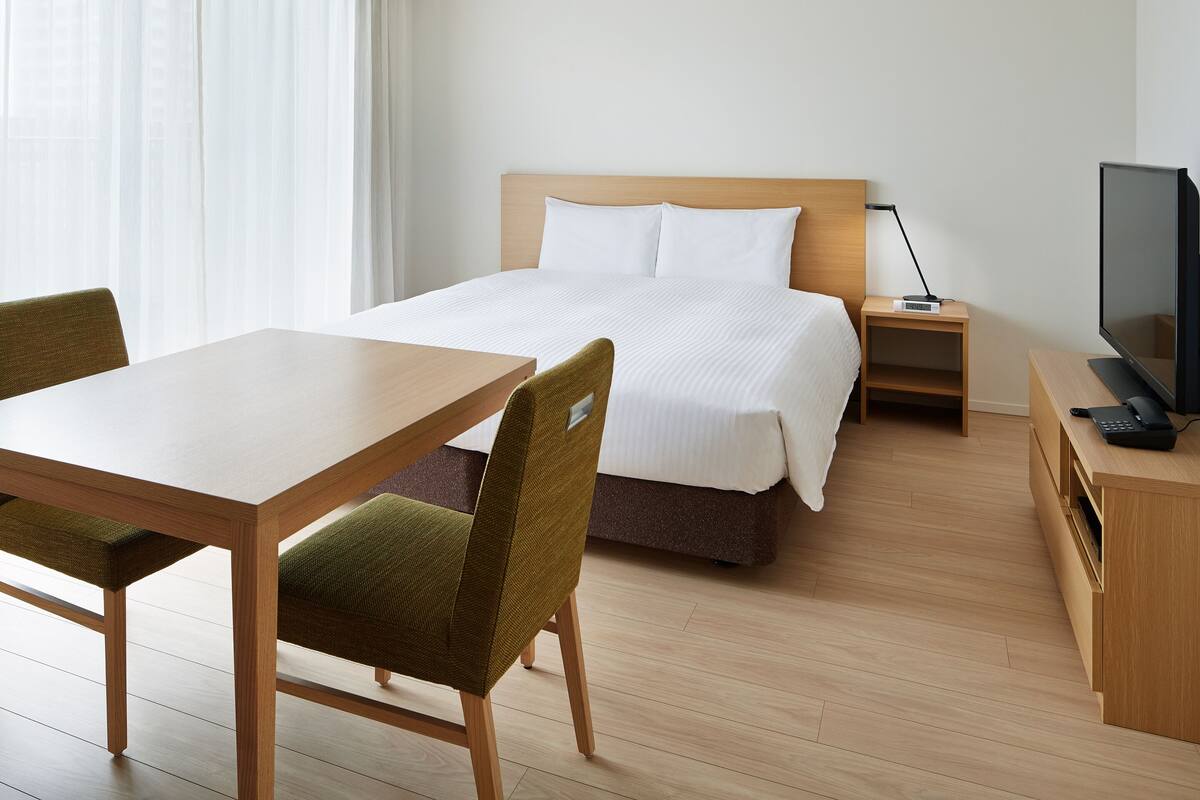 A hotel room with a bed, a desk, a chair, a TV, and a bedside table.