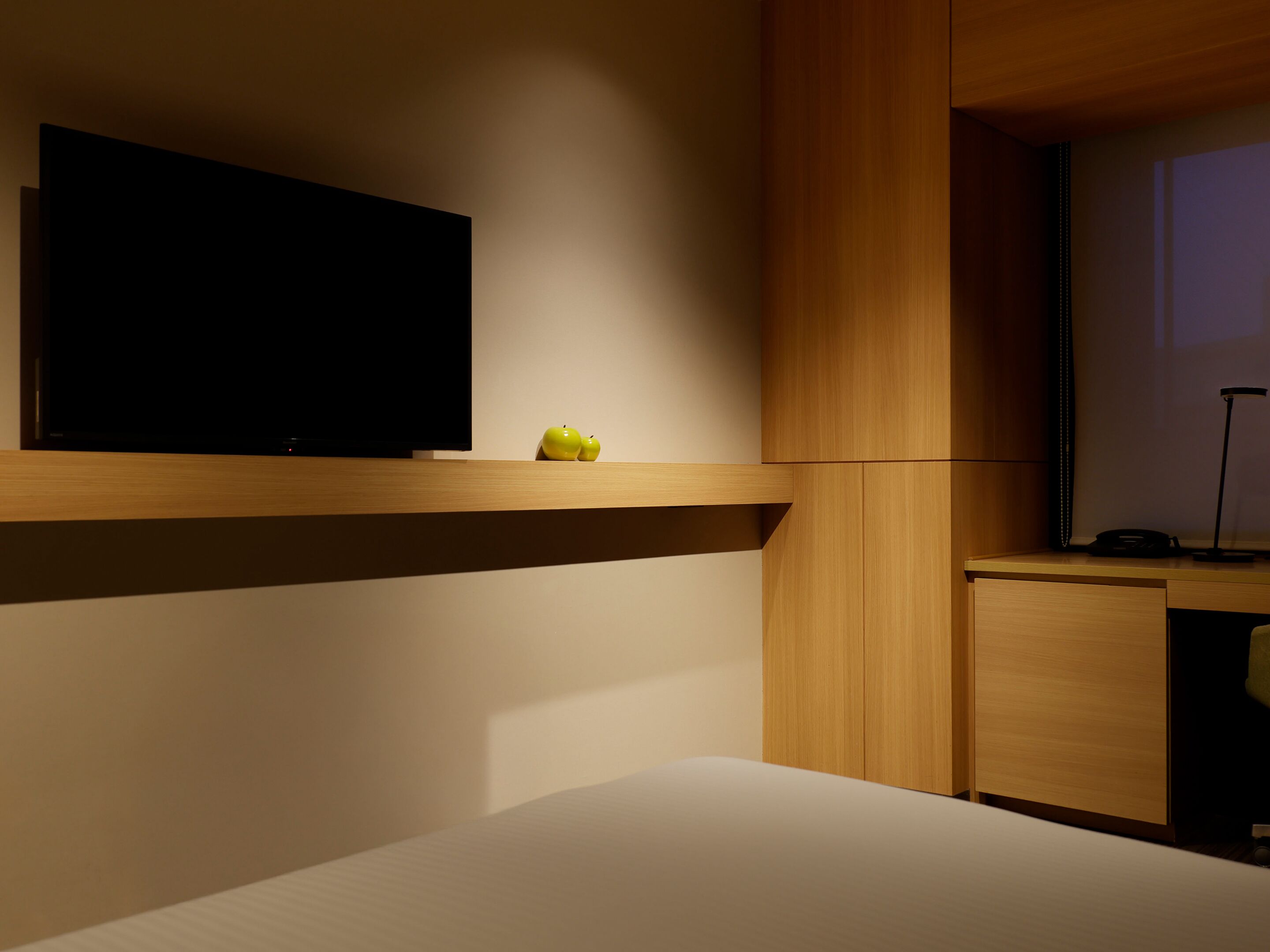 A hotel room with a bed, a flat-screen TV mounted on the wall, a small wooden desk, and a nightstand.
