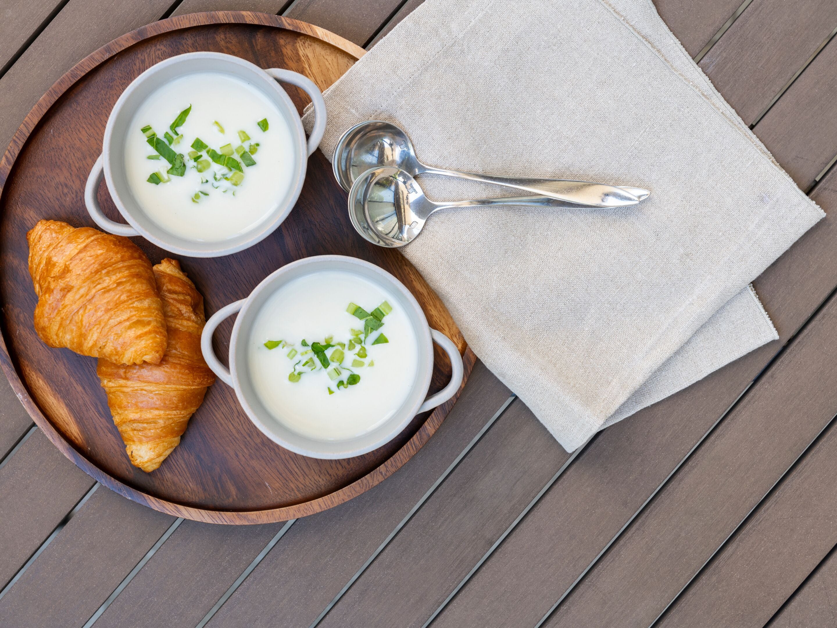 Zwei weiße Suppenschalen mit grüner Dekoration auf einem Holztablett, dazu ein Croissant und ein Silberlöffel.