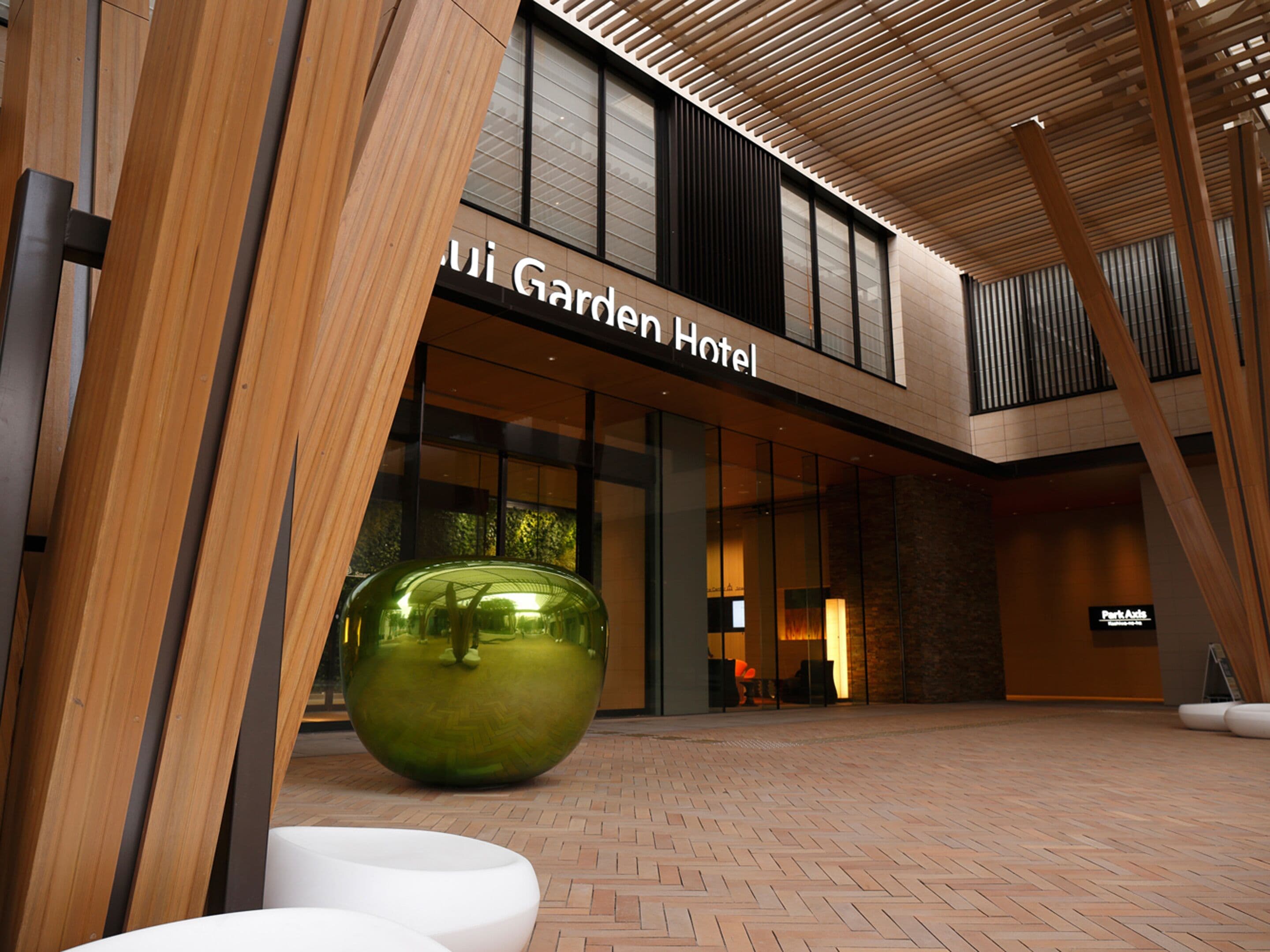 Una entrada de hotel moderna con un amplio frente de vidrio, techo de listones de madera y una llamativa escultura esférica verde al frente.