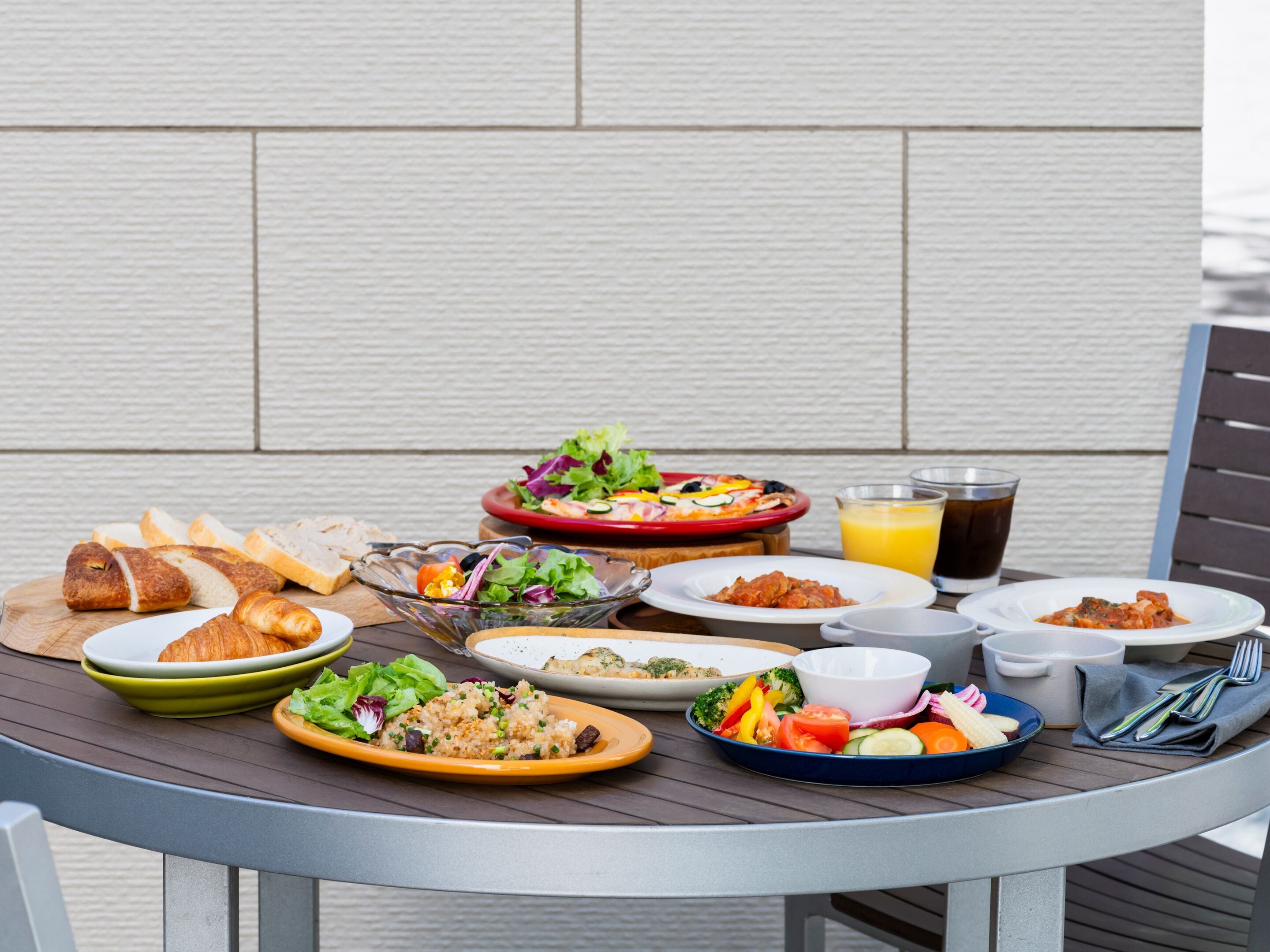 A table set with various dishes including bread, salads, and a pizza, with a drink and utensils on the side.