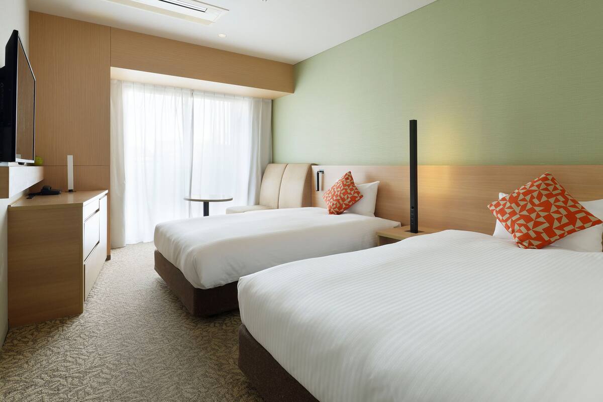 A hotel room with two beds, a desk, a TV, and a window with curtains.