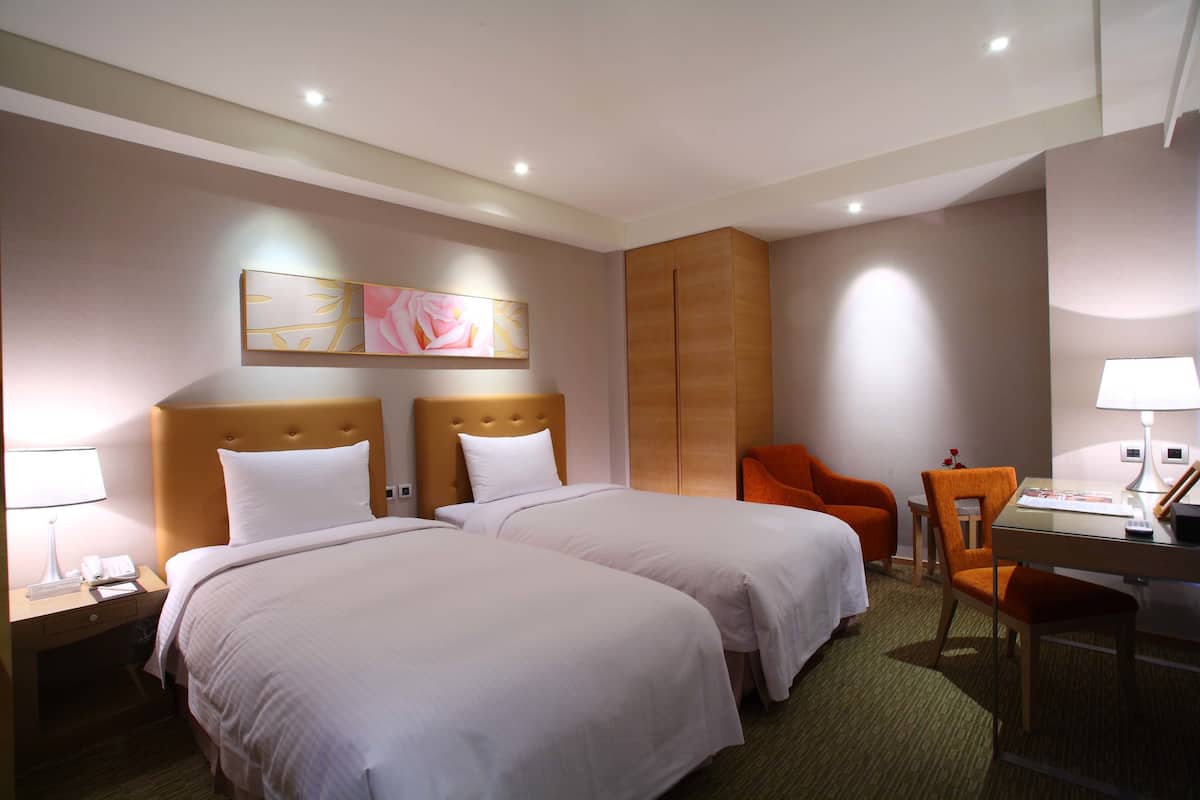 A hotel room with two beds, a desk, a chair, a nightstand, and a lamp.