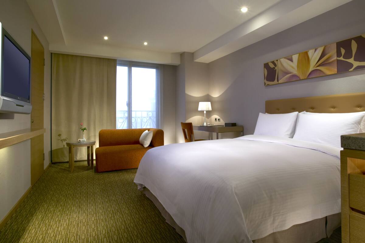 A hotel room with a large bed, a TV mounted on the wall, a desk with a lamp, and a view of the outdoors through a window.
