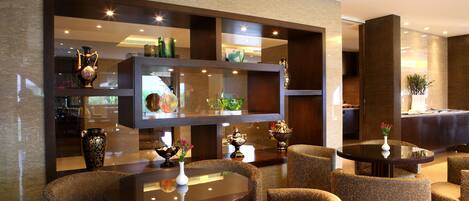 Un vestíbulo moderno con mesas redondas y sillones estampados, una vitrina de cristal con objetos decorativos y un mostrador de recepción.