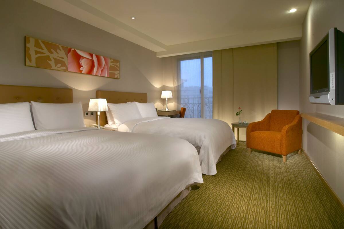 A hotel room with two beds, a small table with a lamp, an orange armchair, a TV, and a balcony with curtains.