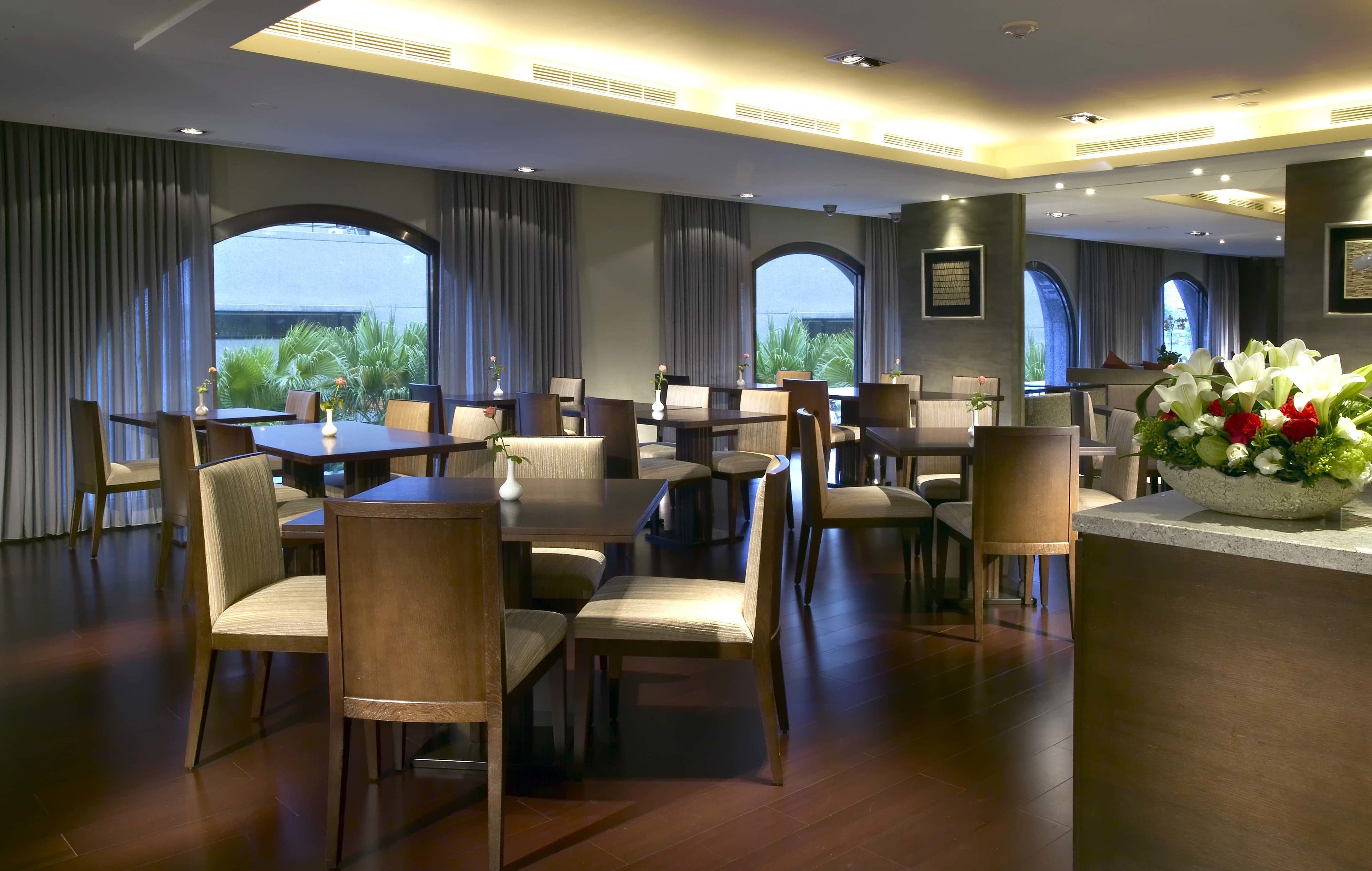 Un comedor de hotel con varias mesas y sillas, amplias ventanas con cortinas y un arreglo floral central.