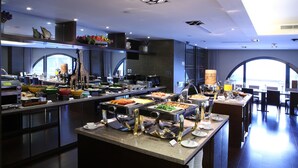 Monipuolinen buffet, jossa on useita ruokapisteitä, kahviautomaatti ja suuri ikkuna, josta avautuu näkymä kaupunkiin.