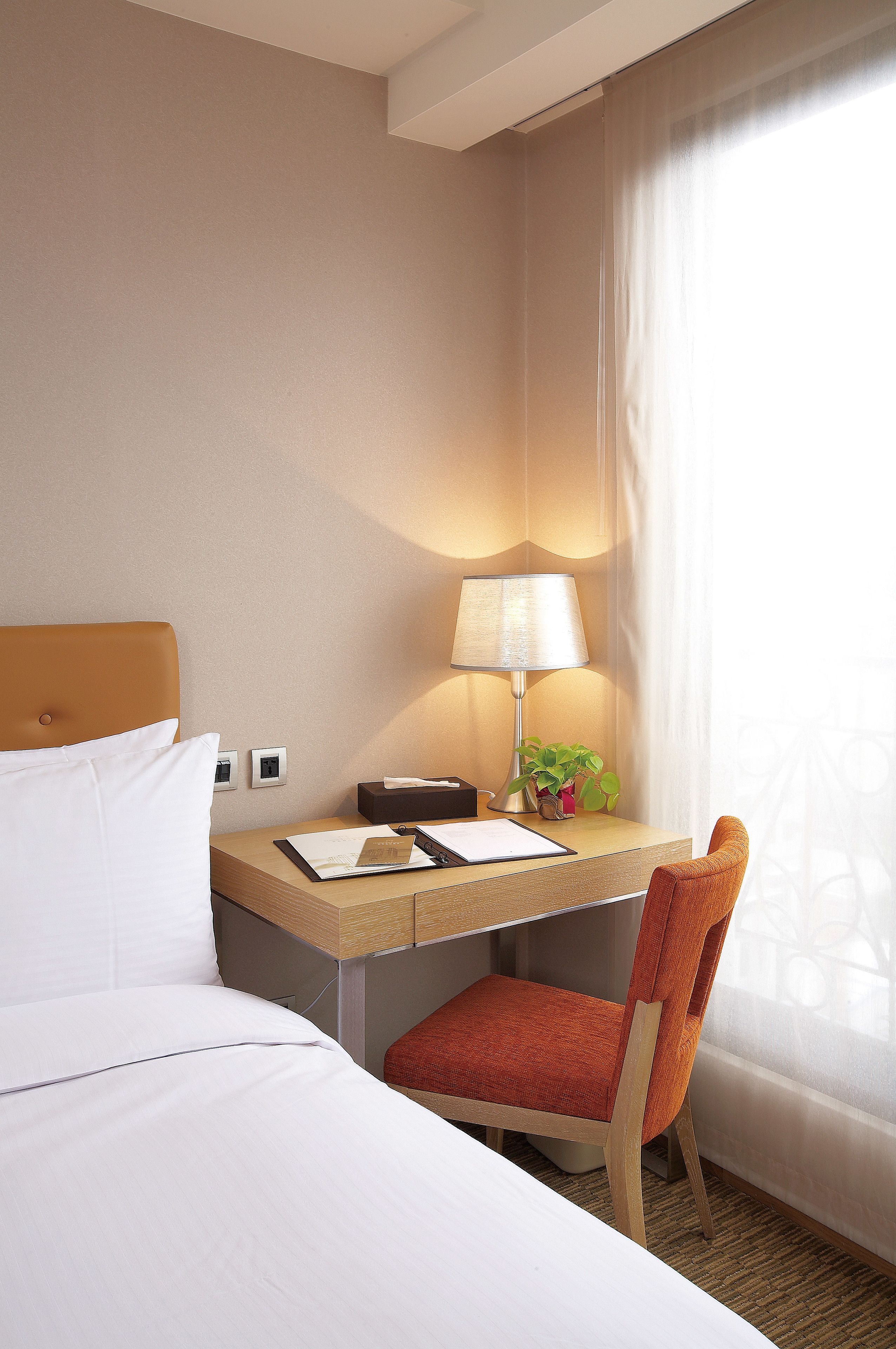Habitación de hotel con cama, escritorio con lámpara, silla y ventana con cortinas.