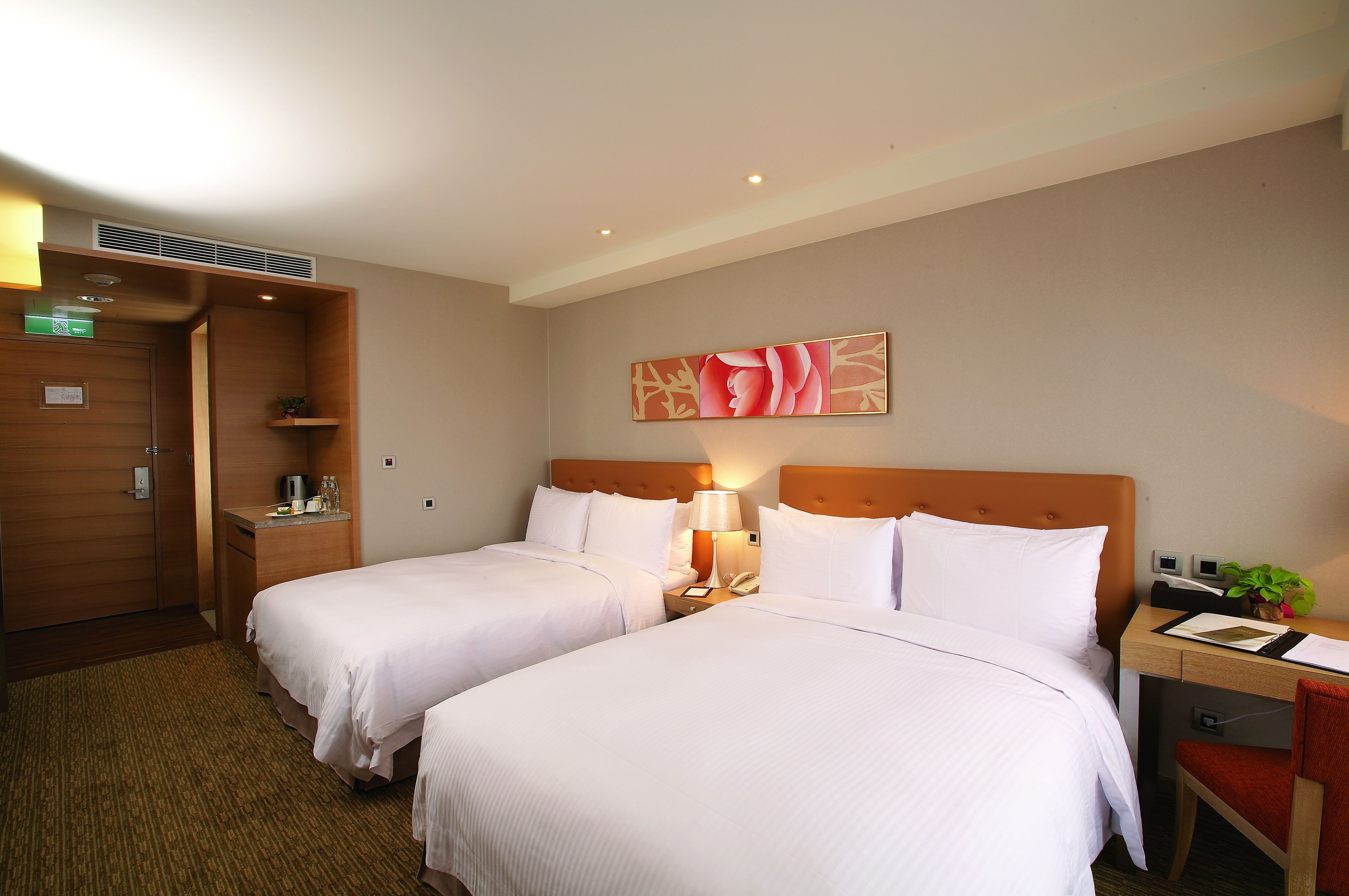 A hotel room with two beds, a wooden wardrobe, a desk with a chair, a lamp, and a wall-mounted artwork.