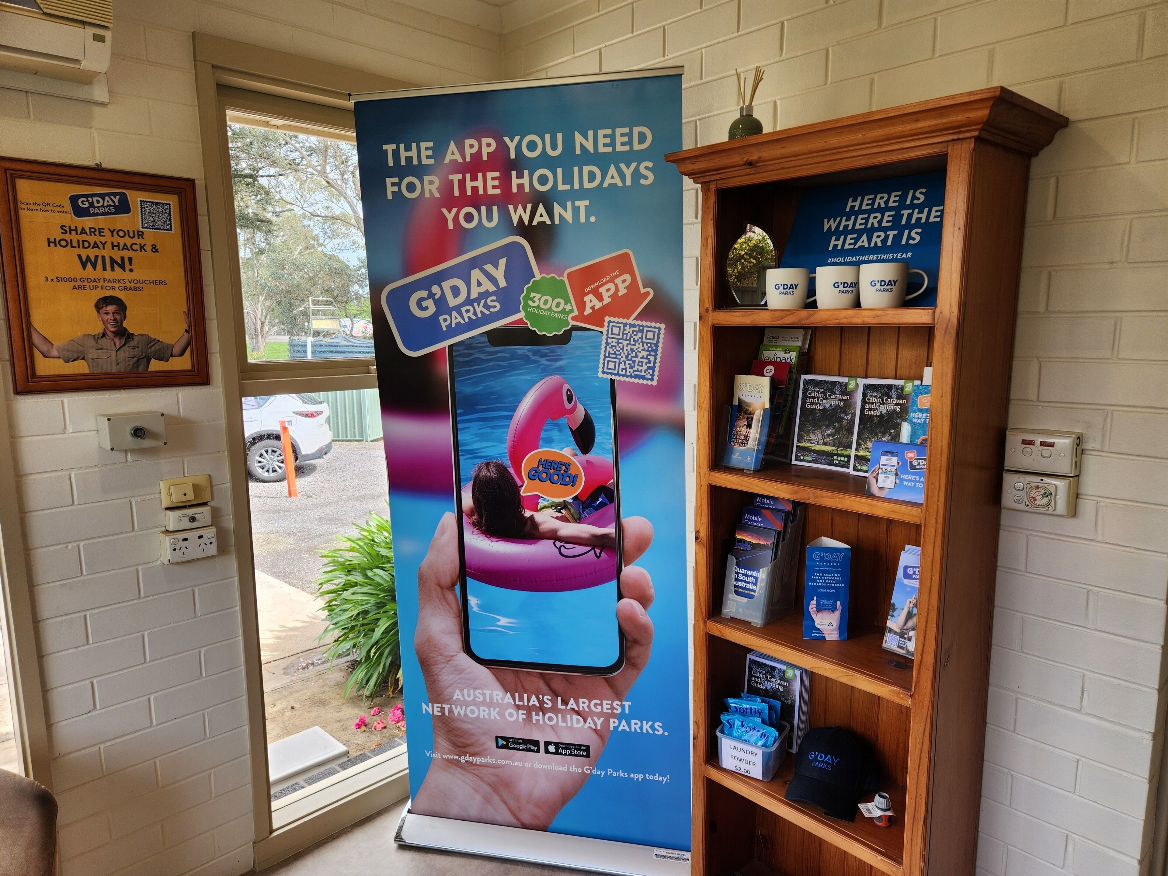 A promotional display for G'DAY Parks featuring a QR code, a G'DAY Parks app advertisement, and a shelf with camping-related items.