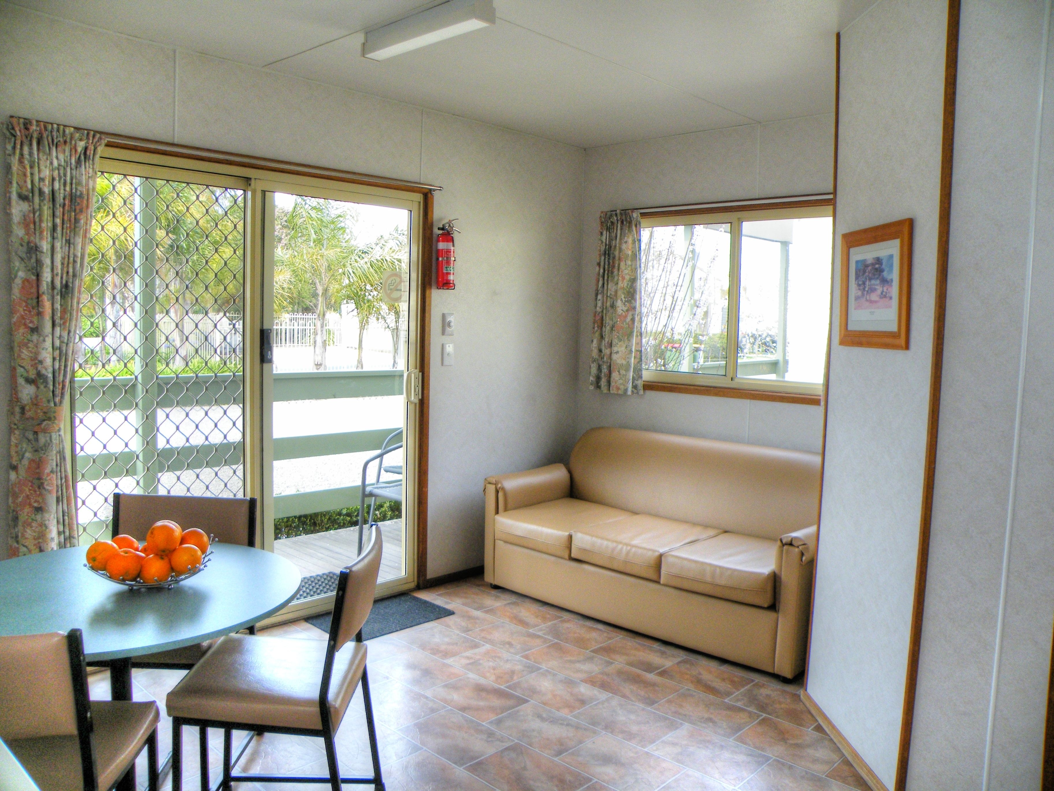 Une pièce avec un canapé beige, une table à manger avec des chaises, une fenêtre avec des rideaux, un extincteur et un tableau encadré au mur.